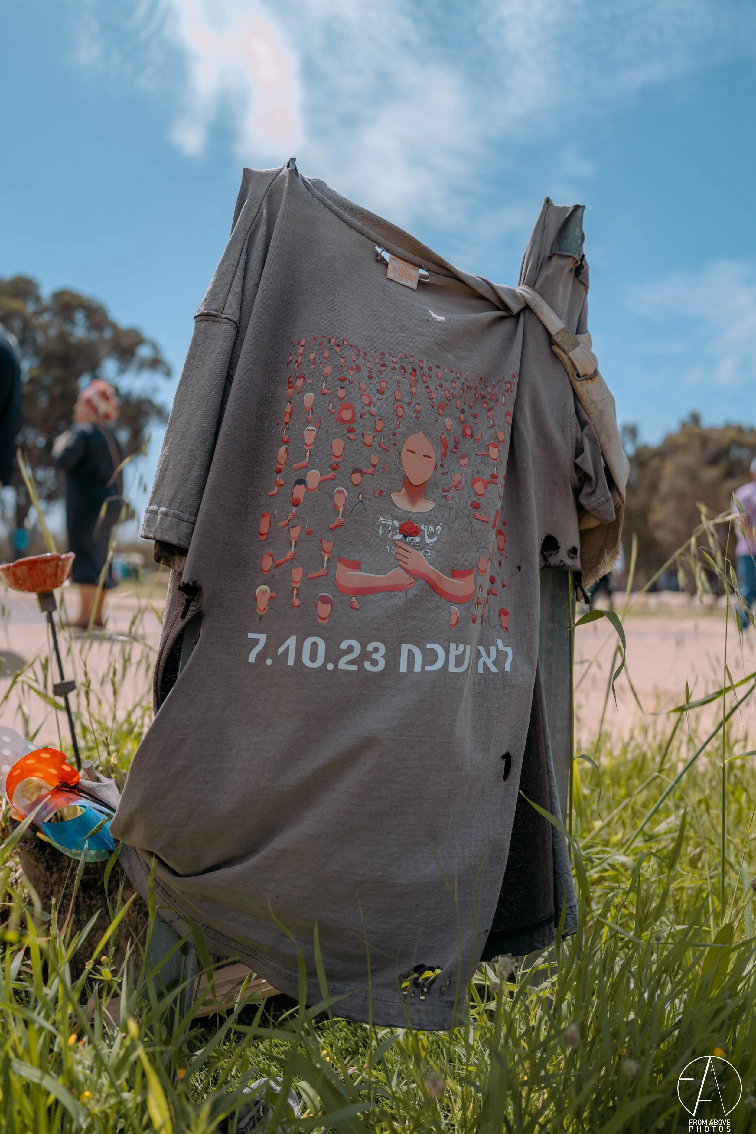 A gray T-shirt hanging outdoors on a grassy field with a design of a person holding a red flower amid other small figures, and Hebrew text with the date 7.10.23 underneath.