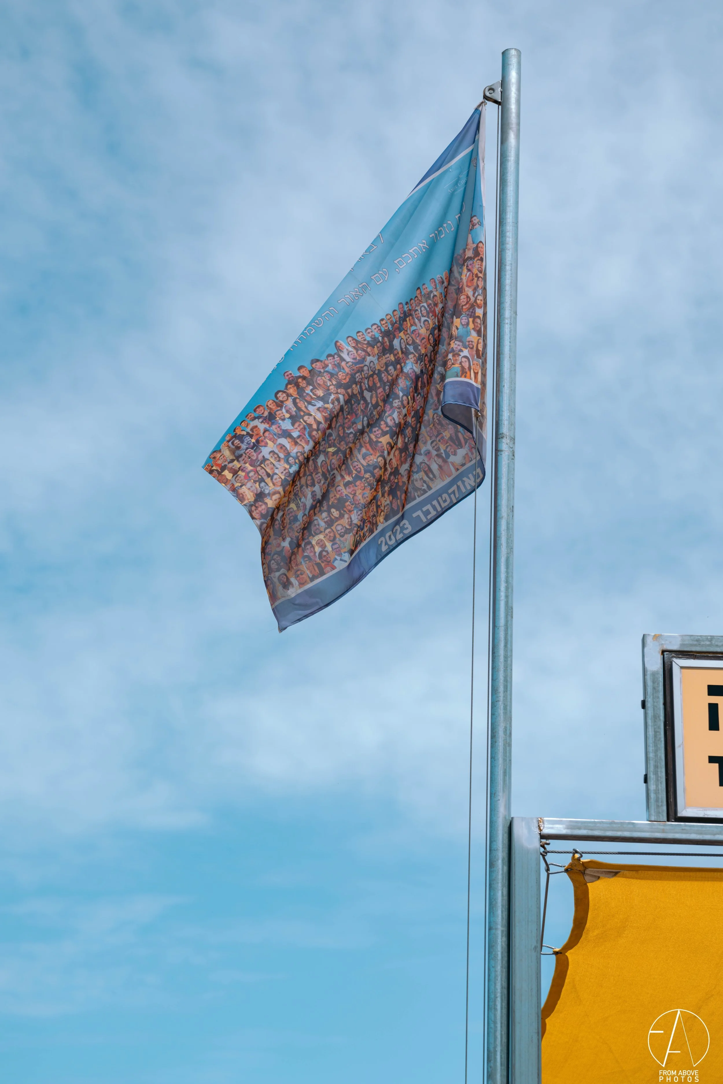 Flag with a group photo of many people, flying against a blue sky, with Hebrew and English text, and yellow part of a sign or banner visible.