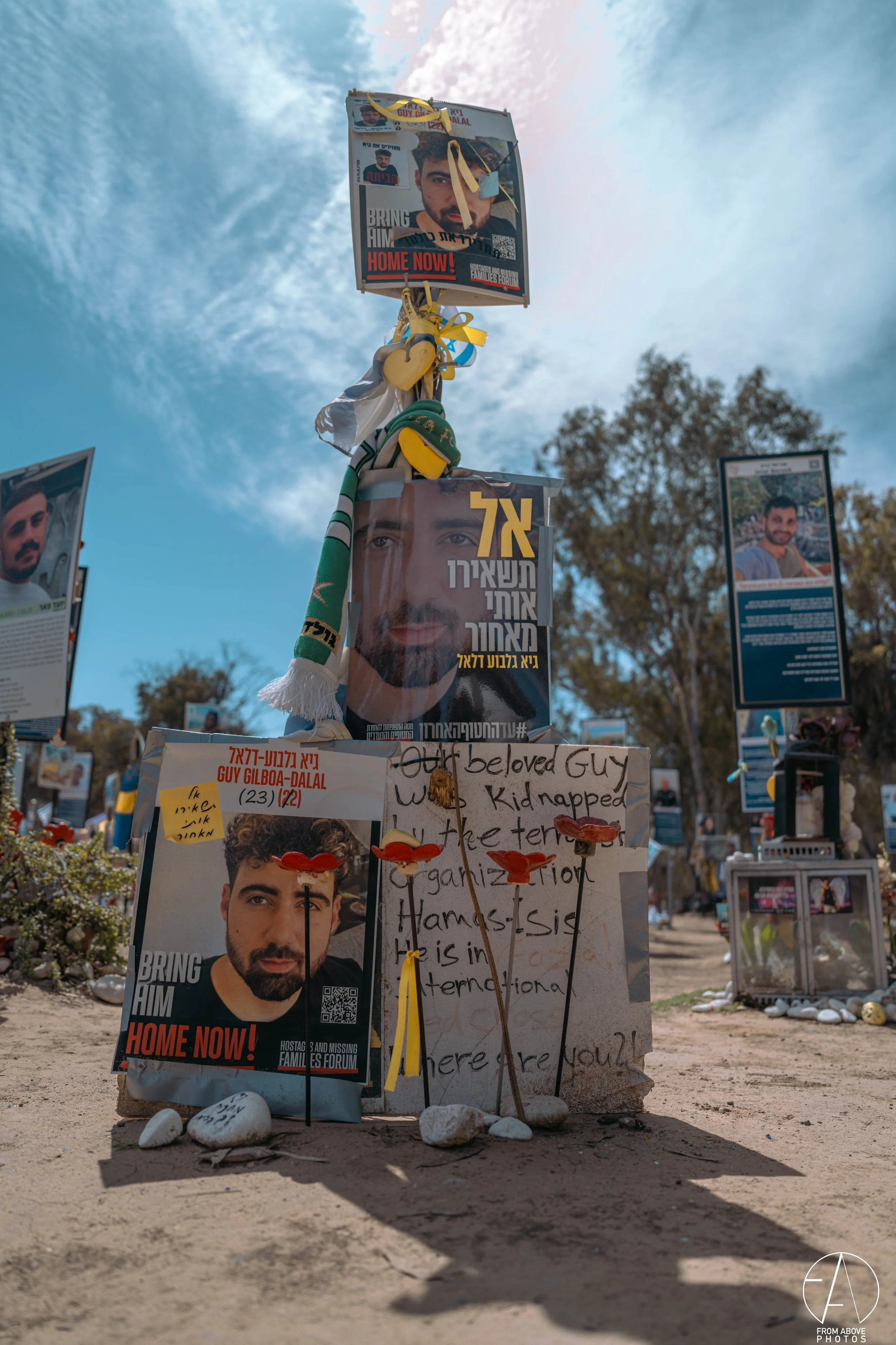 Memorial with posters of a man, ribbons, and notes, set on a stone base surrounded by rocks at an outdoor site with trees and other memorials in the background.
