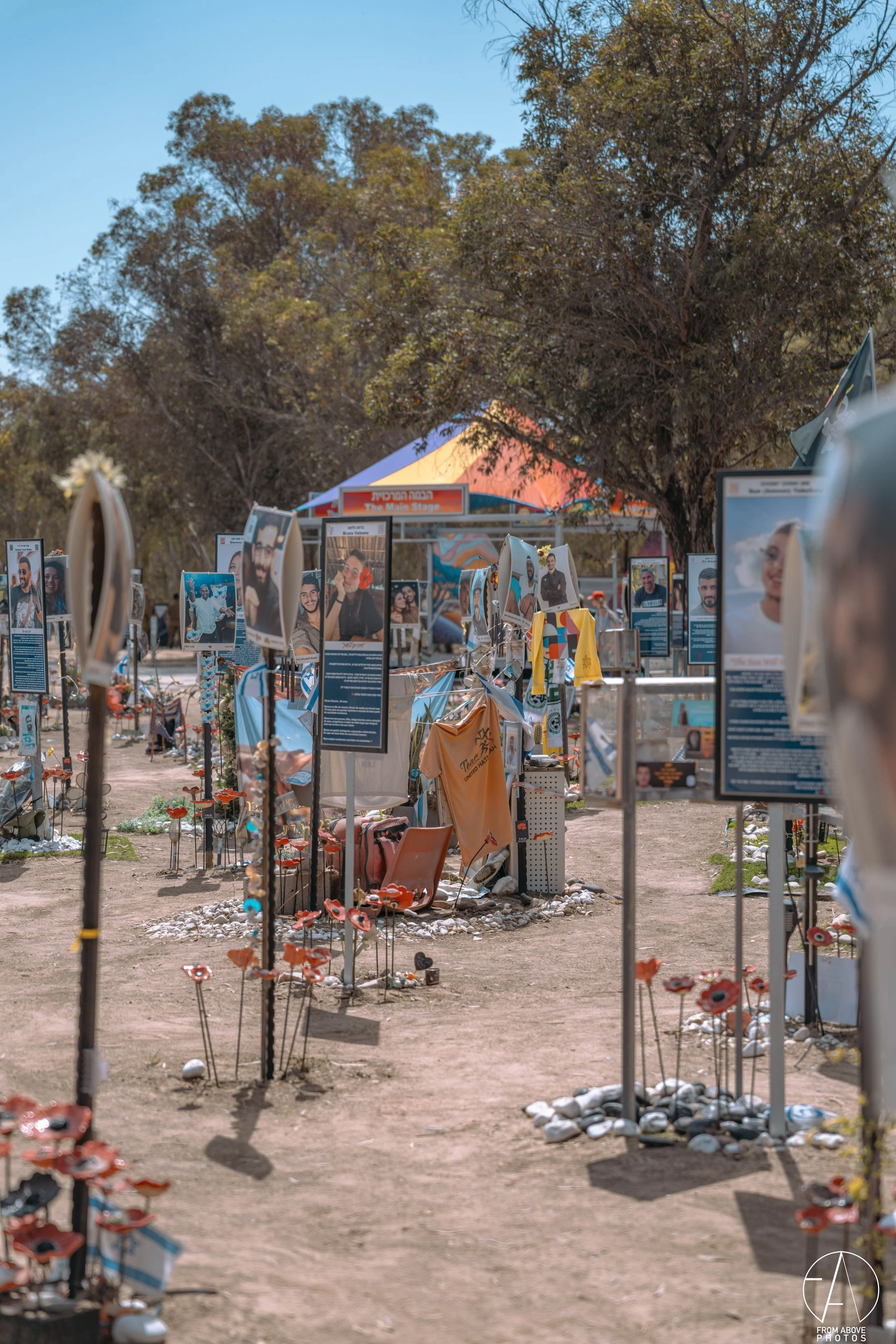 Outdoor memorial display with photographs, flowers, and personal items under trees, with a tent in the background.