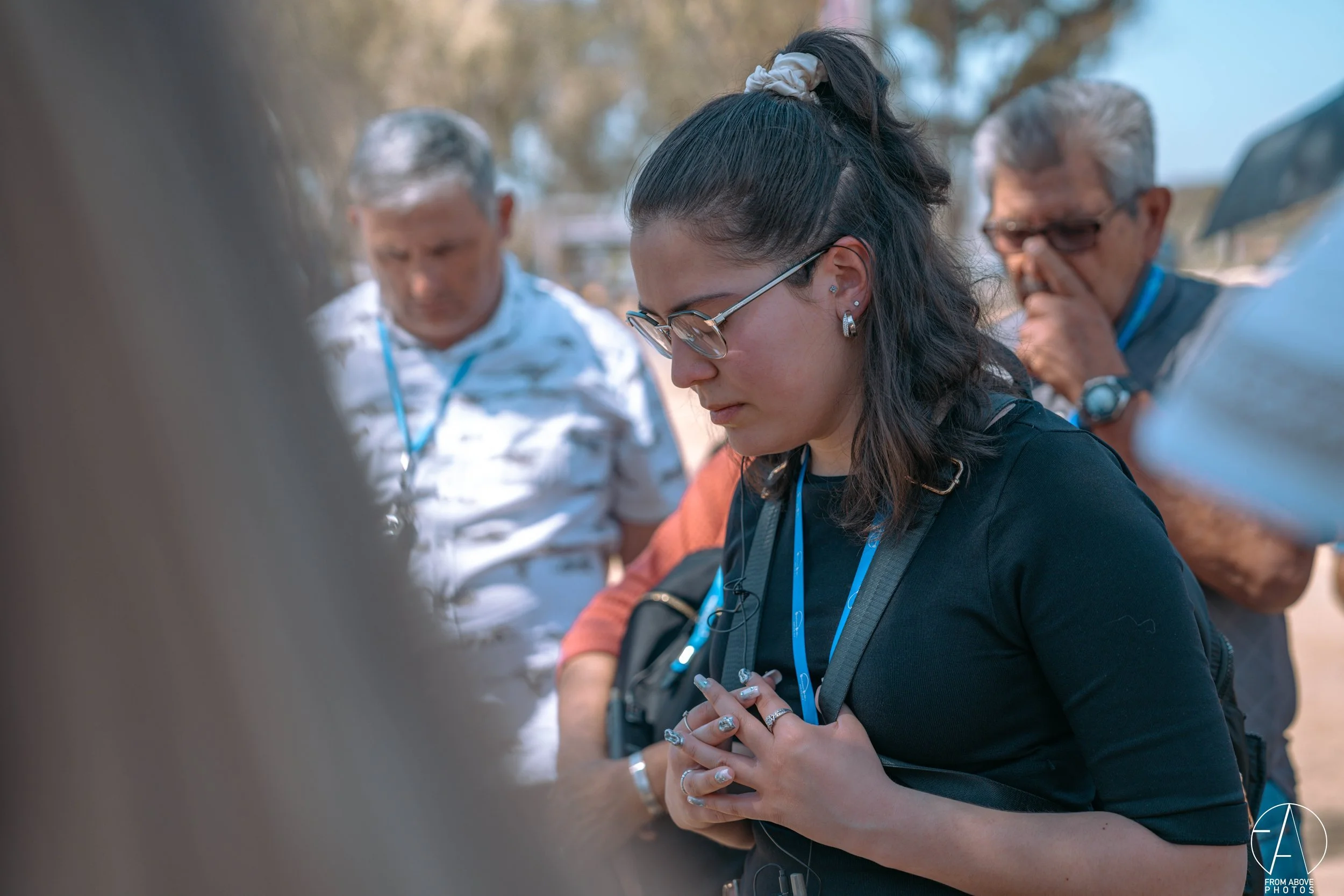 A young woman with glasses and dark hair tied back in a ponytail, praying with her hands clasped together, surrounded by a group of people outdoors.