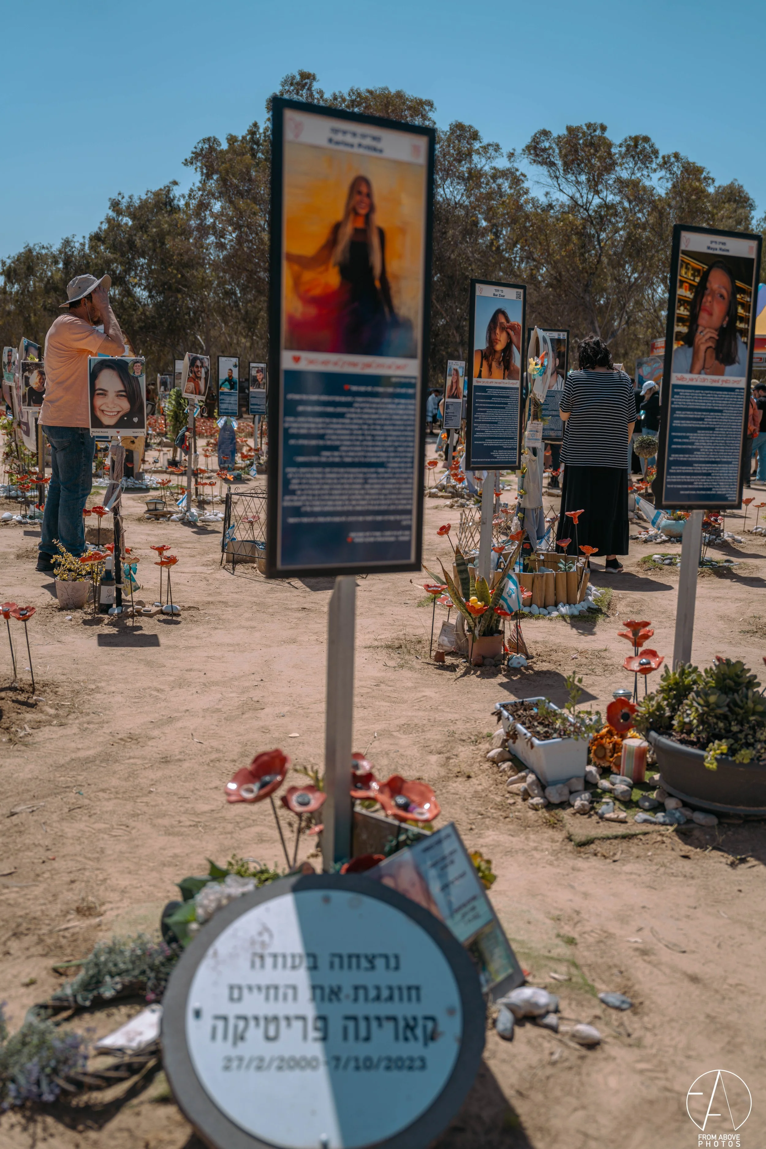 A memorial funeral tribute with photos of a woman and others, surrounded by flowers and candles, in an outdoor setting with trees and a clear blue sky.