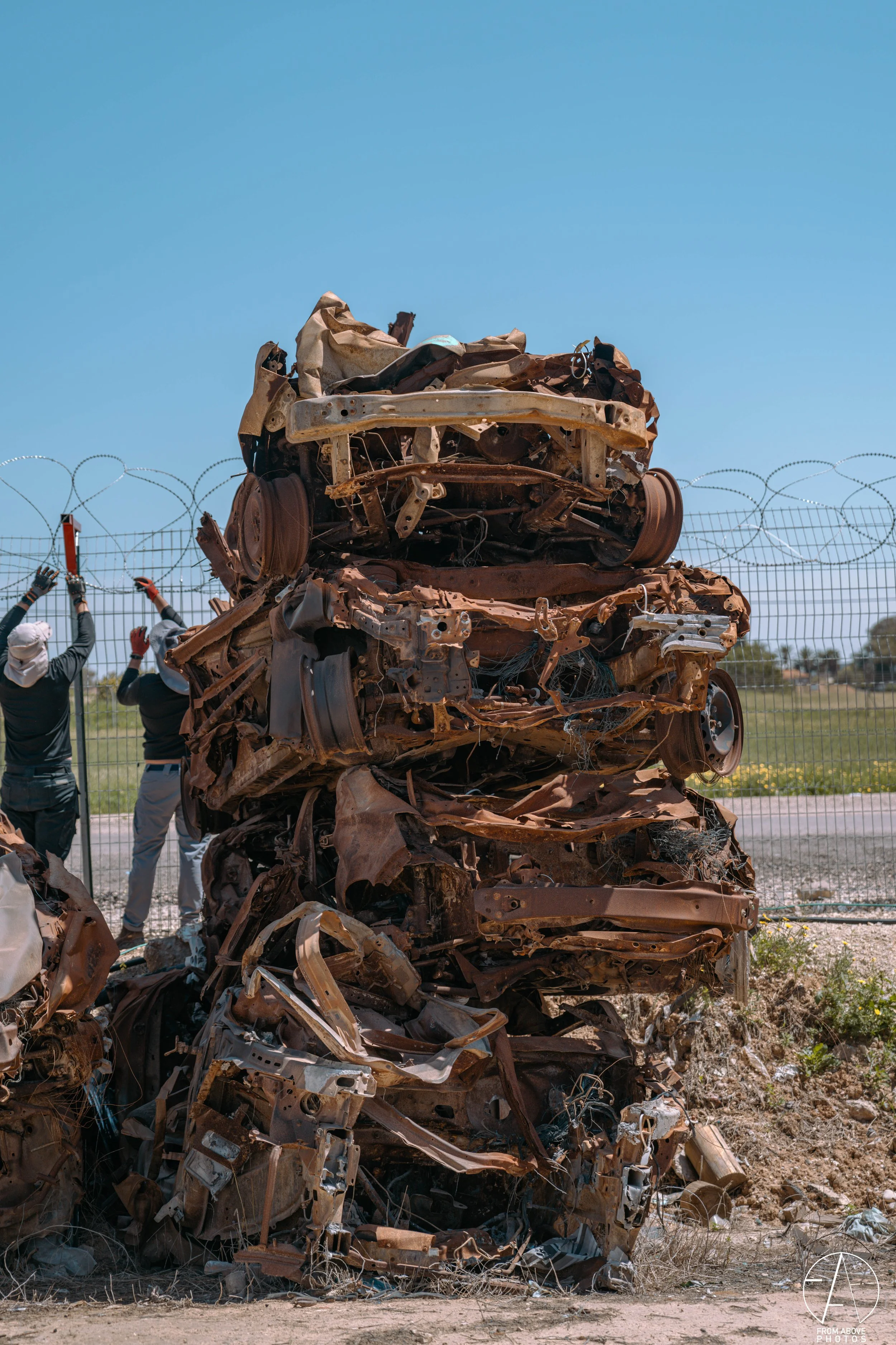 Piles of crushed and twisted wrecked cars stacked outdoors near a barbed wire fence under a clear blue sky.