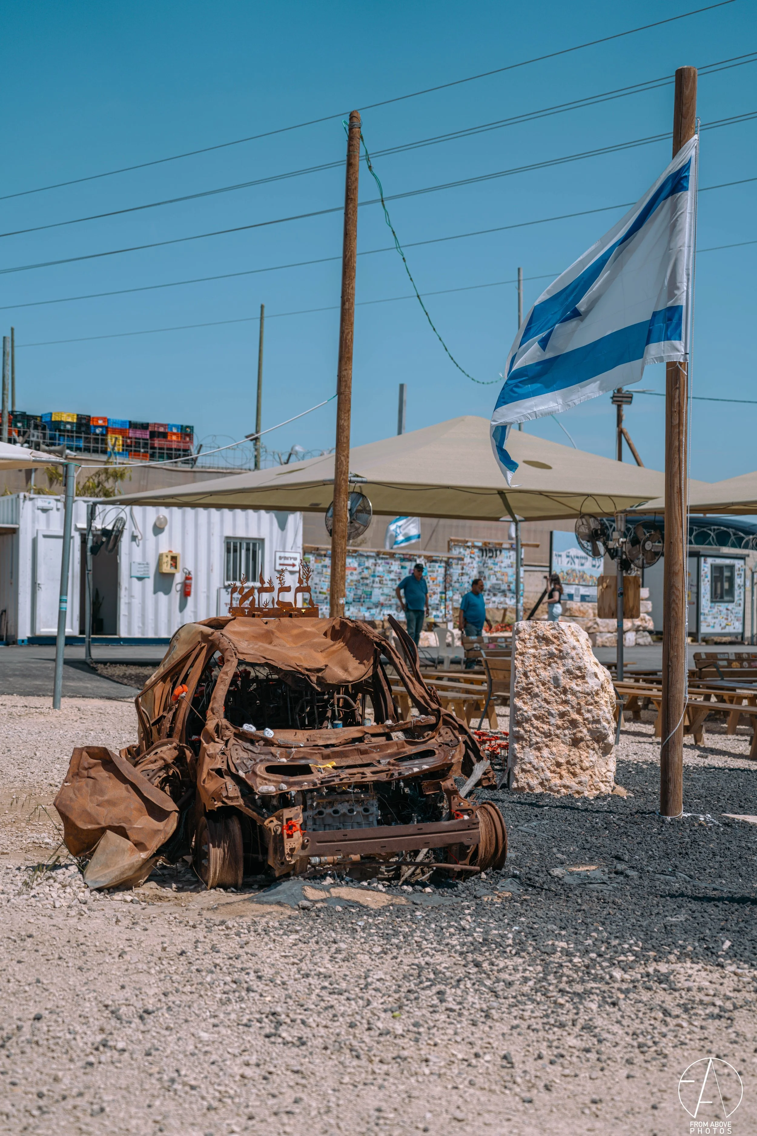 Wreckage of a destroyed car on gravel ground outdoors, with a large stone and a flagpole nearby, under a clear blue sky. There are people in the background near a seating area and a building with outdoor decorations.