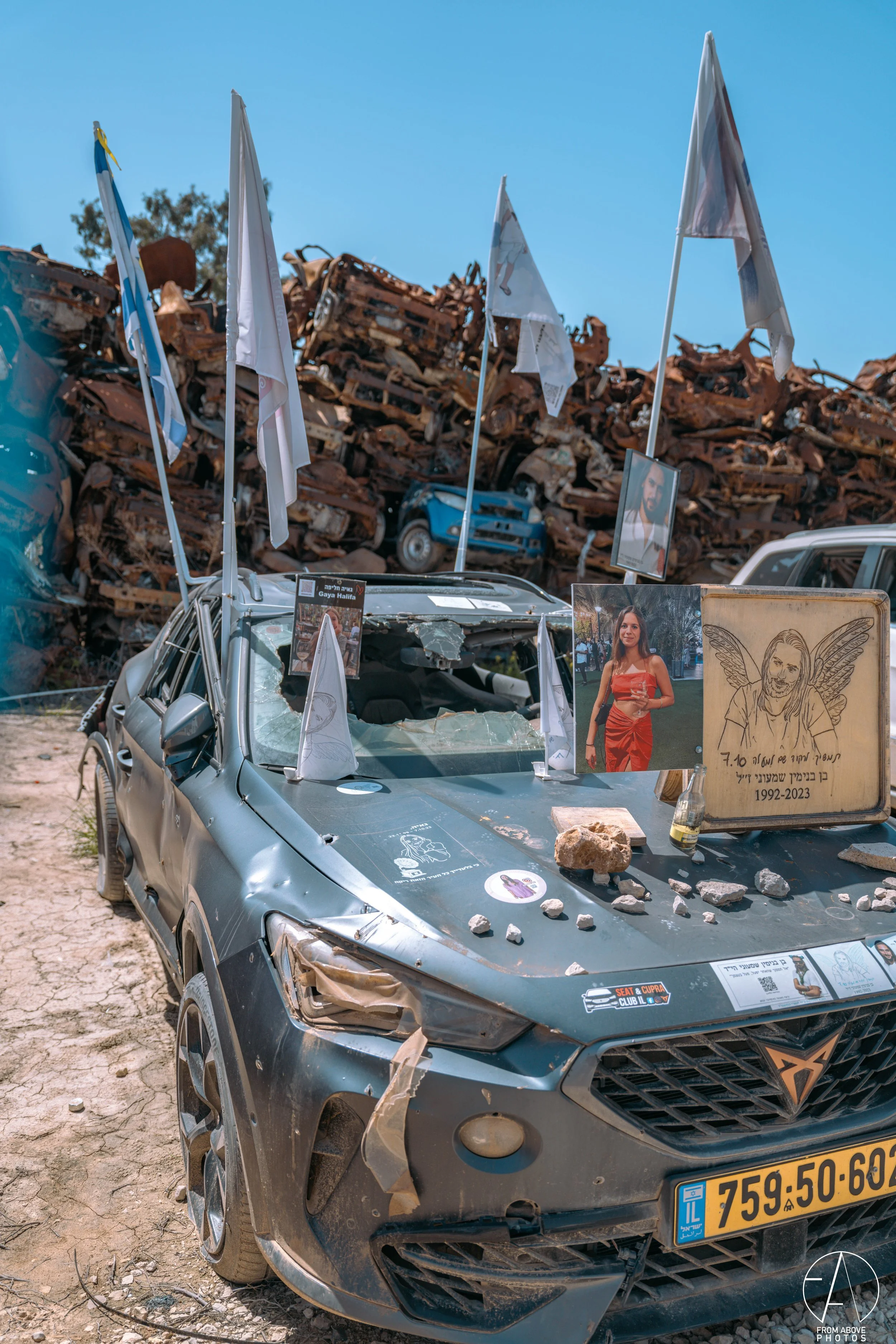 Damaged car at a memorial site with photos, flags, and rocks on the hood, with a pile of scrap metal in the background.