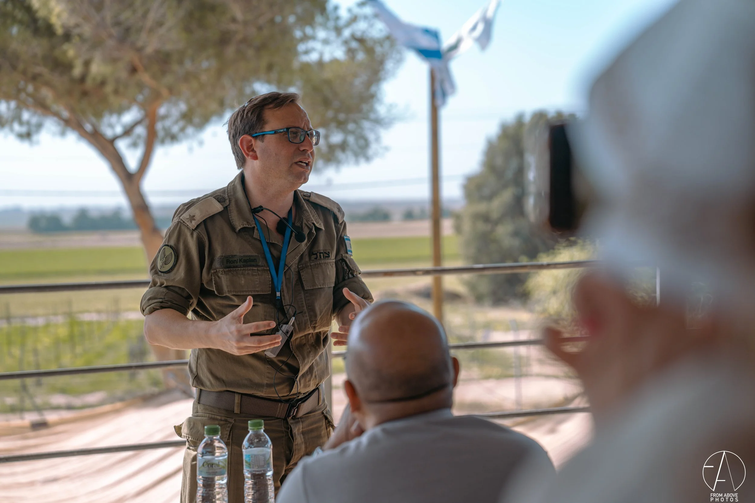 A man in military uniform giving a presentation outdoors with a landscape background, two water bottles on the table, and a few people in the audience.