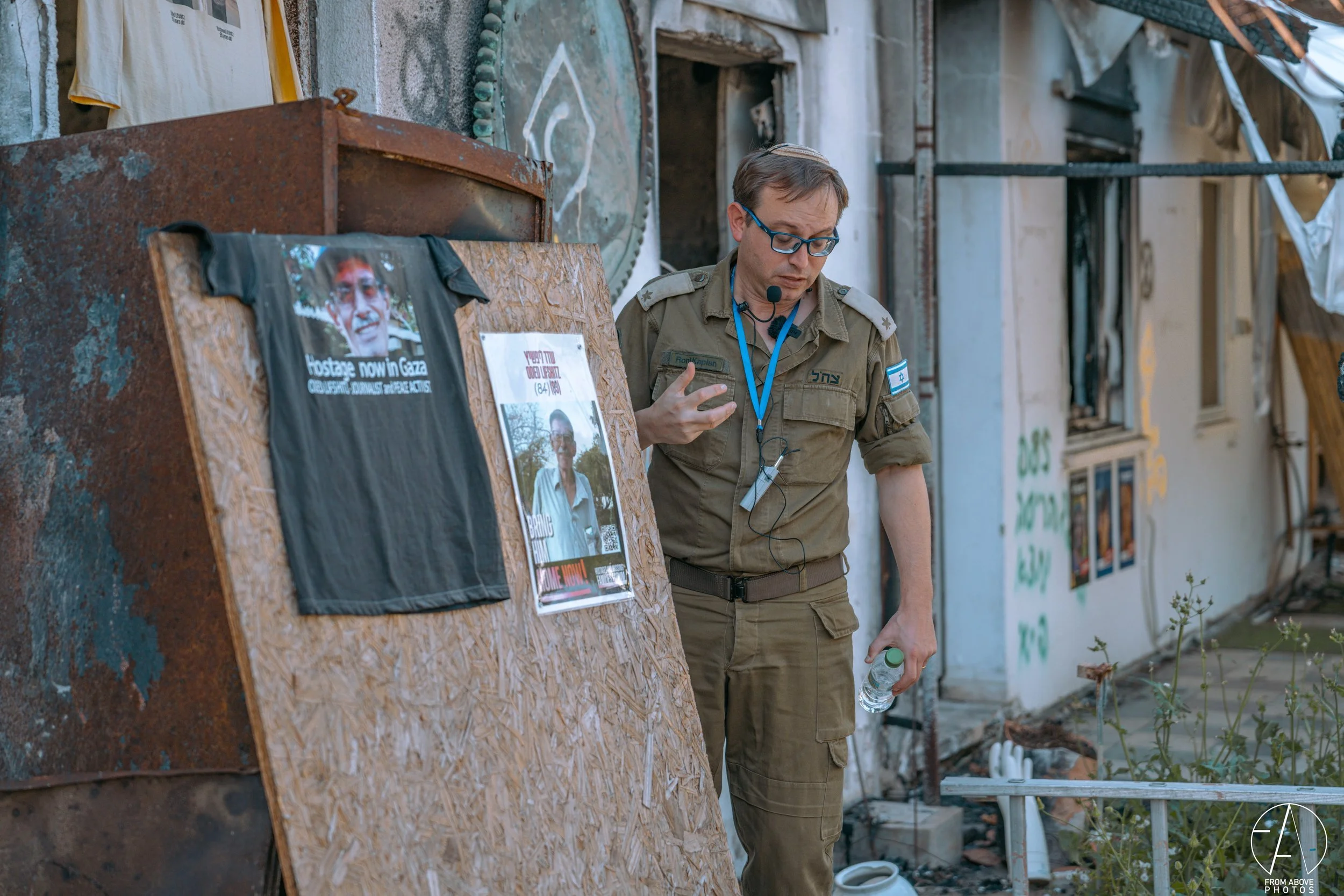 Military personnel standing beside a wooden display board with posters and a shirt, in an outdoor setting with damaged buildings and graffiti.