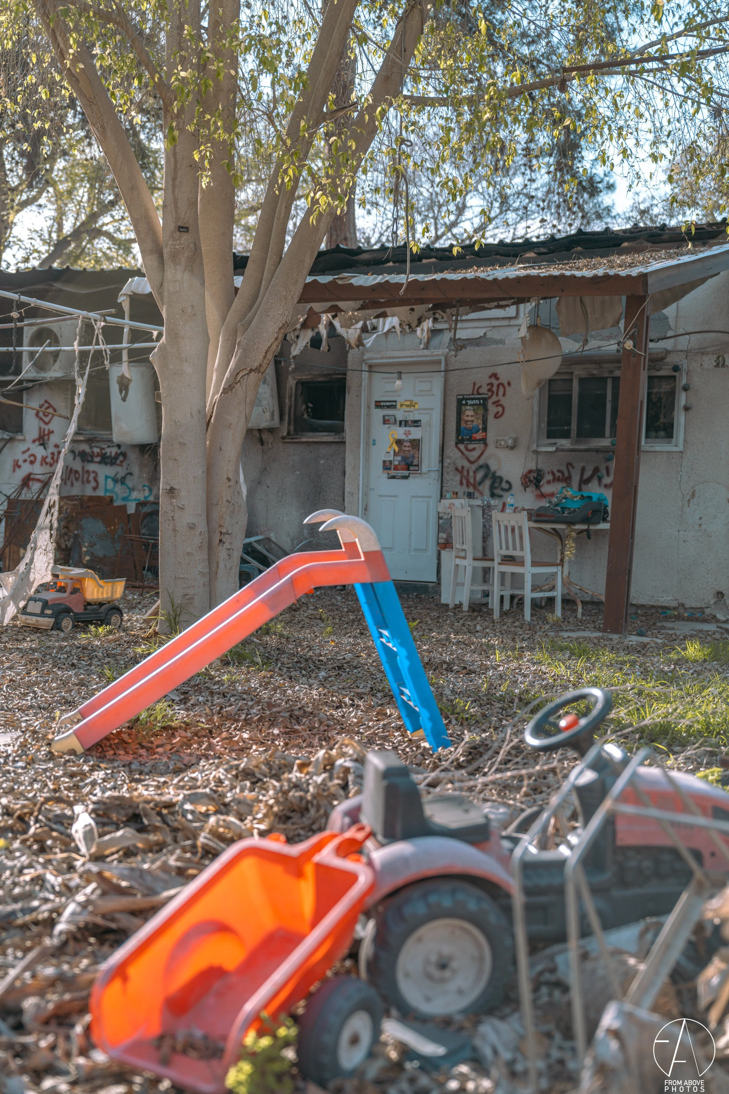 A dilapidated house with graffiti on the walls, broken windows, and a sagging porch. Children's toys including a slide, toy car, and wheelbarrow are scattered on the ground outside, which is covered with leaves.