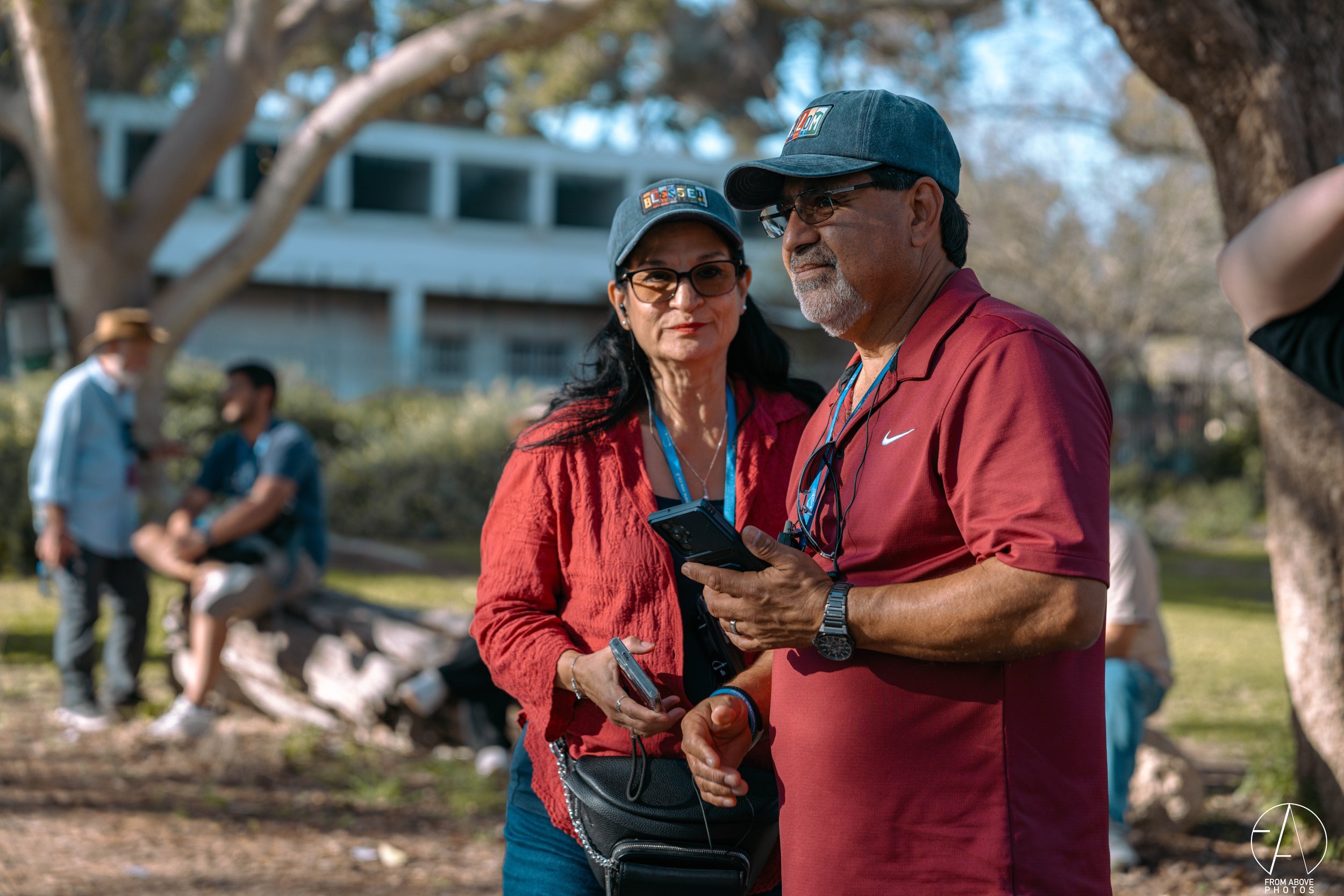 A woman and a man standing outdoors in a park, looking at a cell phone together. The woman is wearing a red jacket and glasses, and the man is wearing a red polo shirt, glasses, and a blue cap. They are under a tree with other people in the backgroun