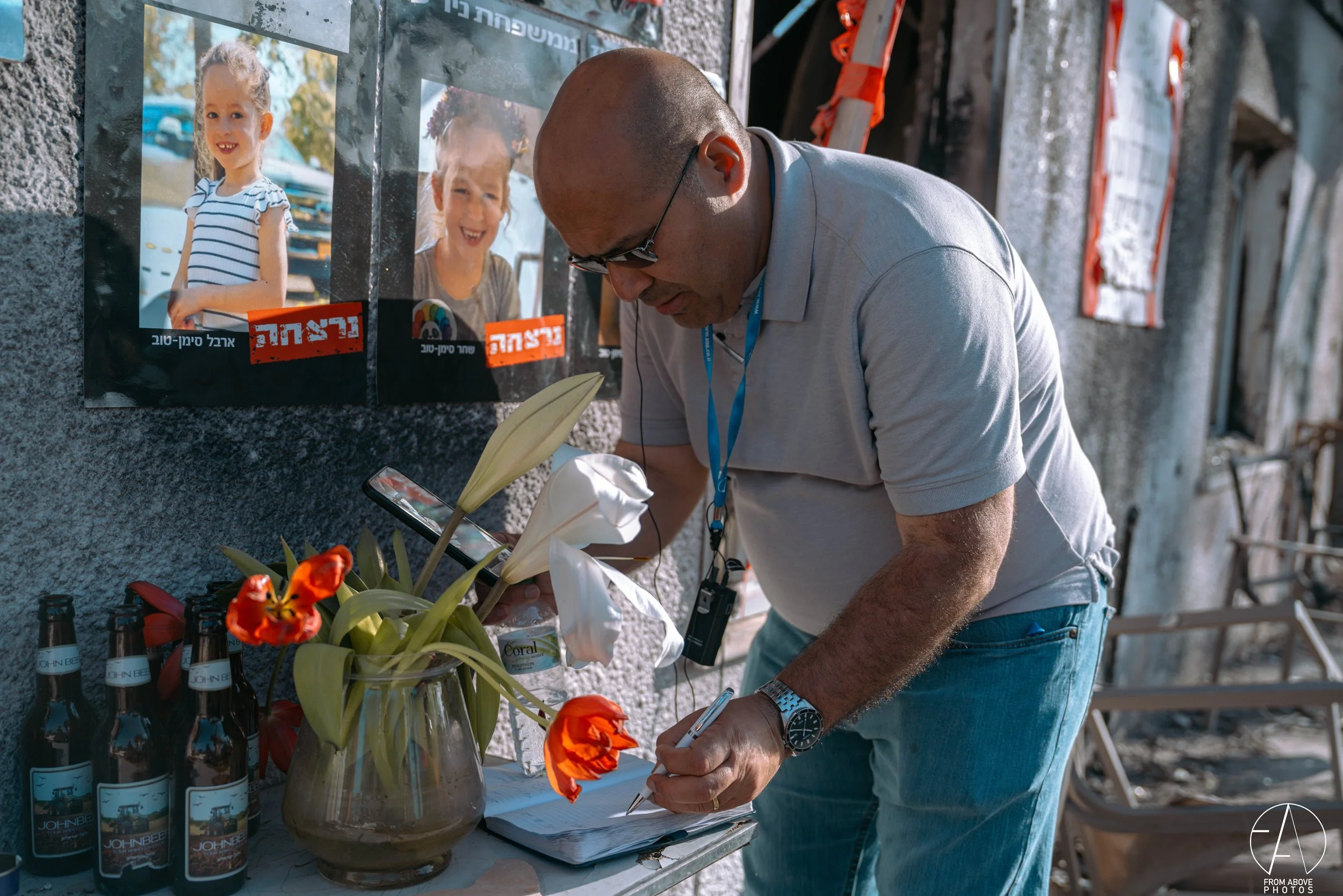 A man wearing glasses and a light gray polo shirt, with a blue lanyard around his neck, is writing in a notebook on a table. The table has a vase with white lilies and orange flowers, several bottles of a beverage labeled 'John B', and a poster with 