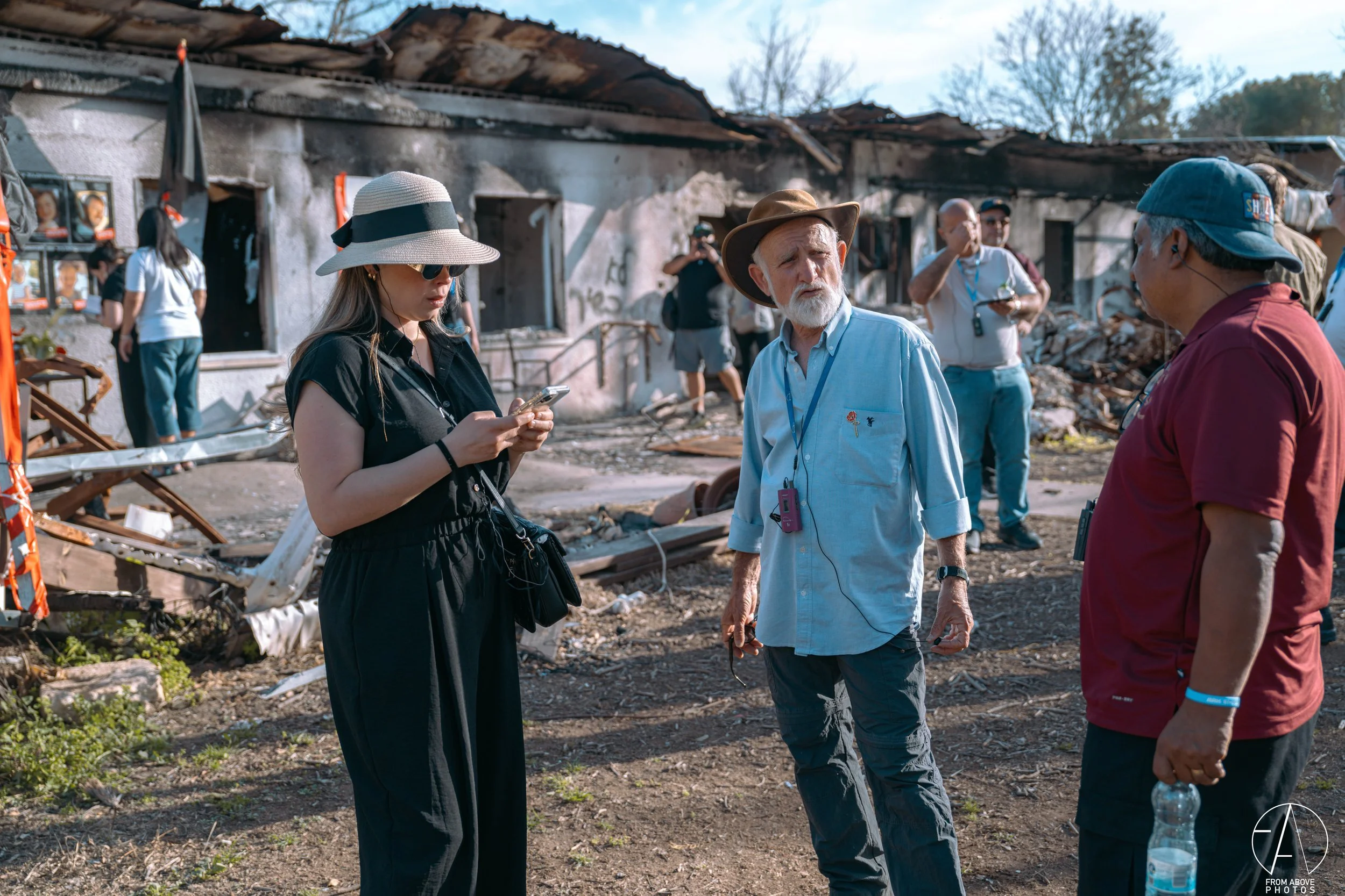 Group of people standing in front of a burned building, discussing the damage. One woman is looking at her phone, while others are chatting, with debris and damaged structure in the background.