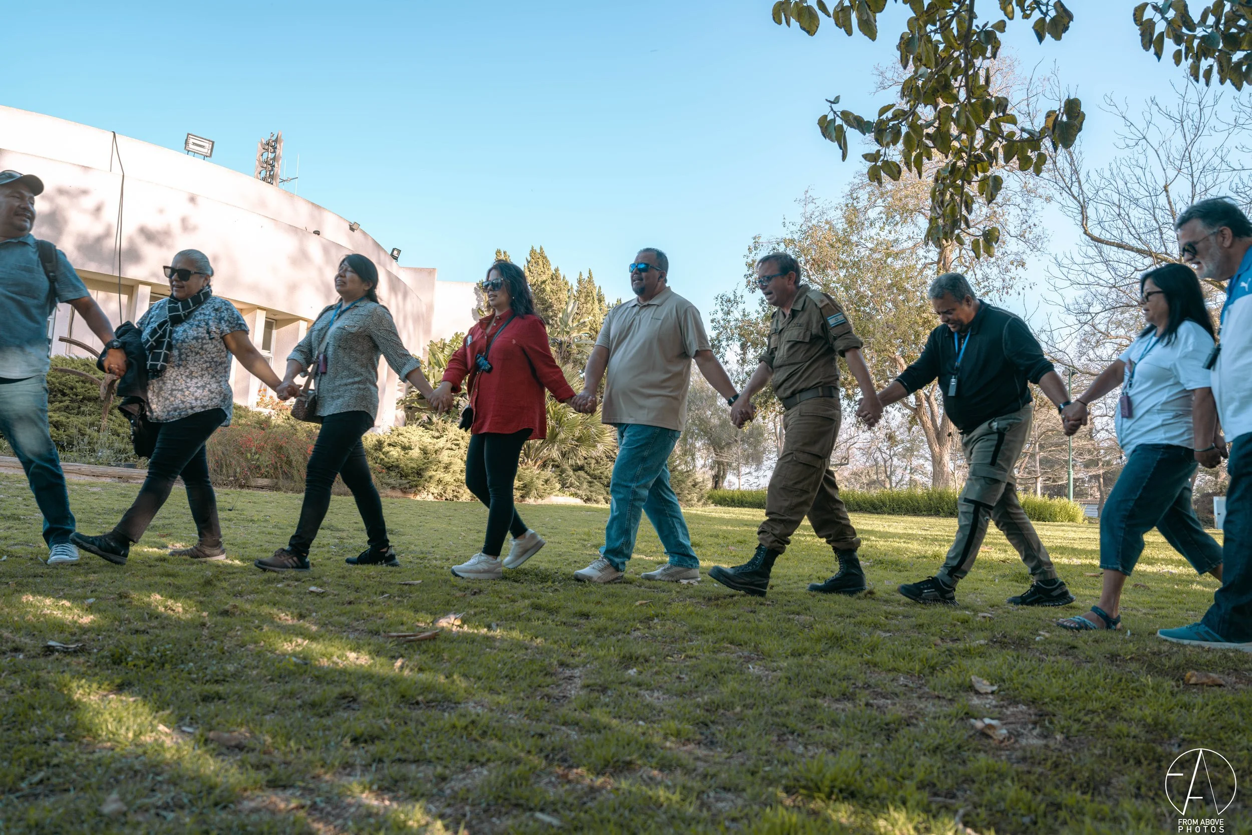 A group of diverse people holding hands in a circle on a grassy outdoor area, outdoors with trees and a white building in the background, under clear blue sky.