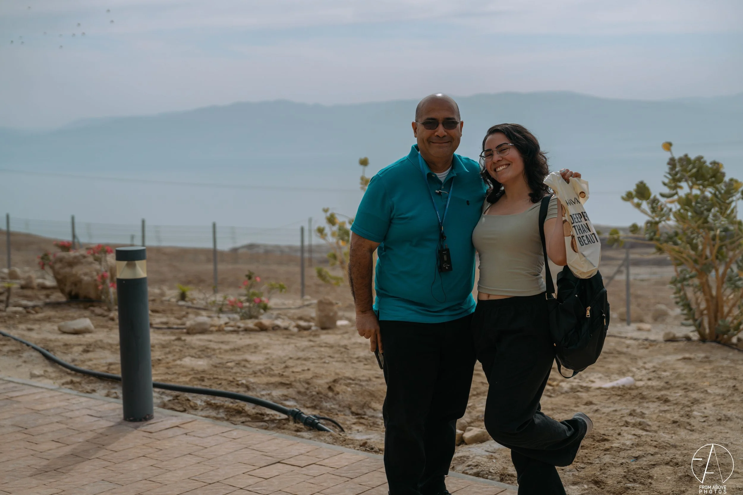 A man and a woman smiling and posing together outdoors on a rocky, desert-like terrain with mountains and a body of water in the background. The man is wearing sunglasses, a turquoise polo shirt, and dark pants, while the woman has glasses, a gray to