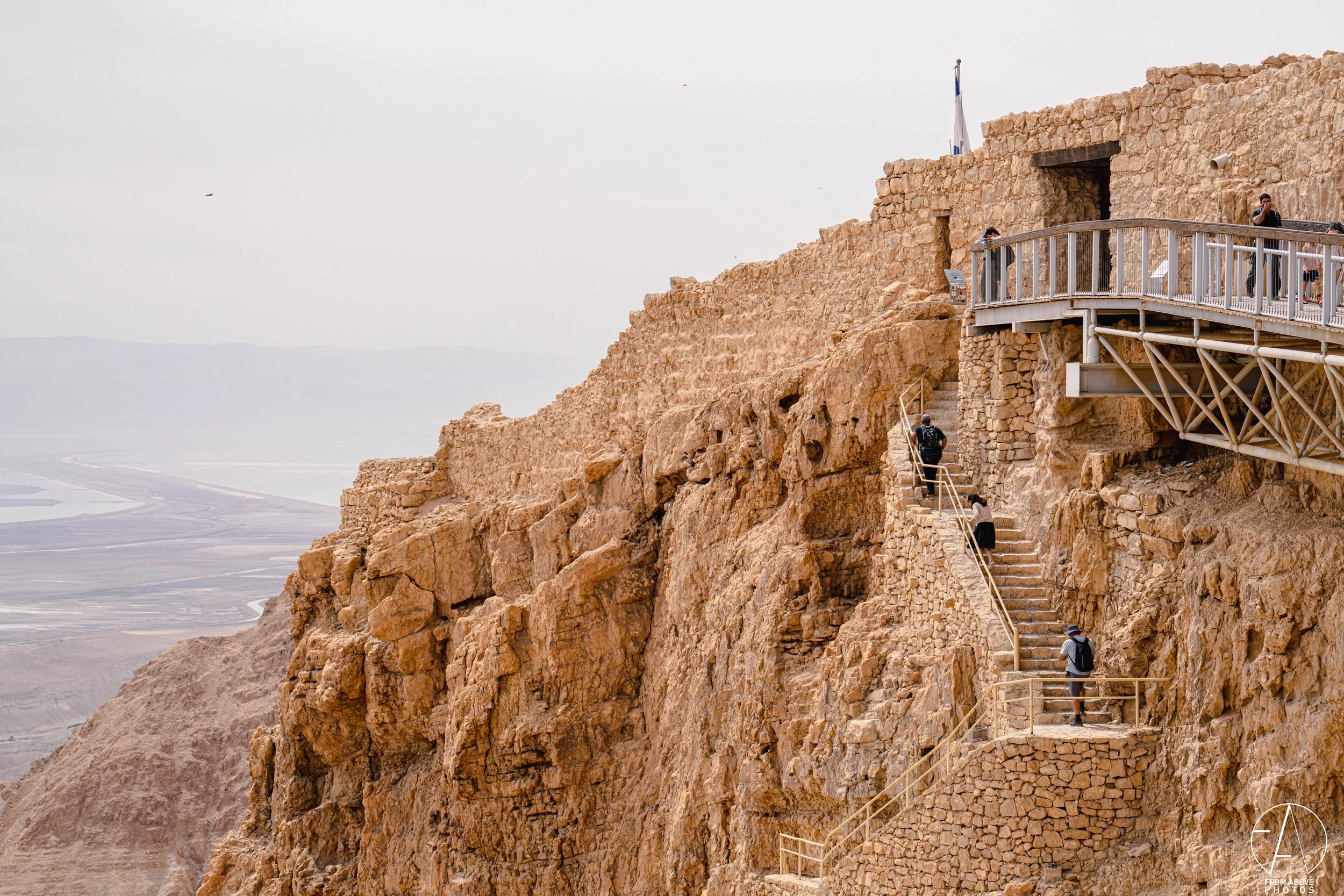 Tourists walking up a staircase along a rugged, rocky cliffside with a stone fortress on top and a viewing platform to the right, overlooking a vast desert landscape.