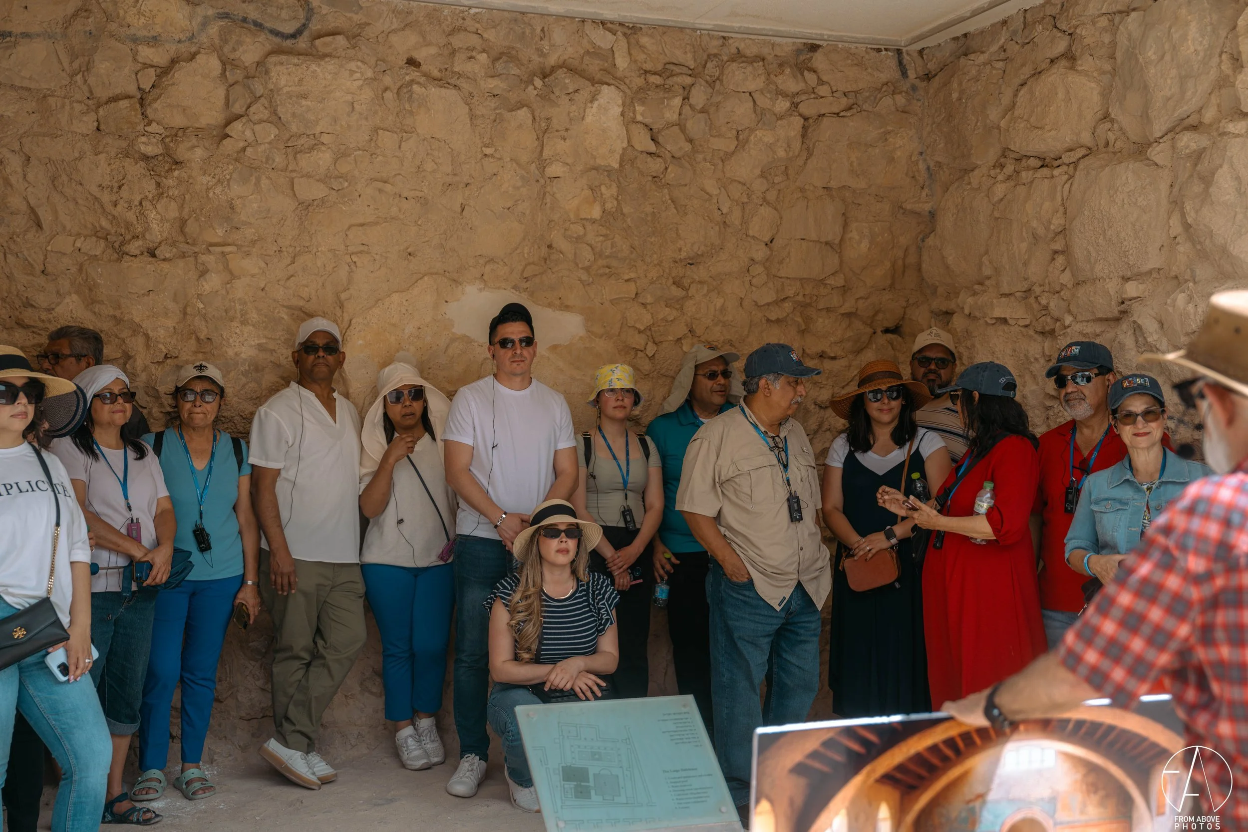Group of tourists on a guided tour inside an archaeological or historical site, standing against a stone wall, some wearing hats and sunglasses, with a tour guide partially visible in the foreground.