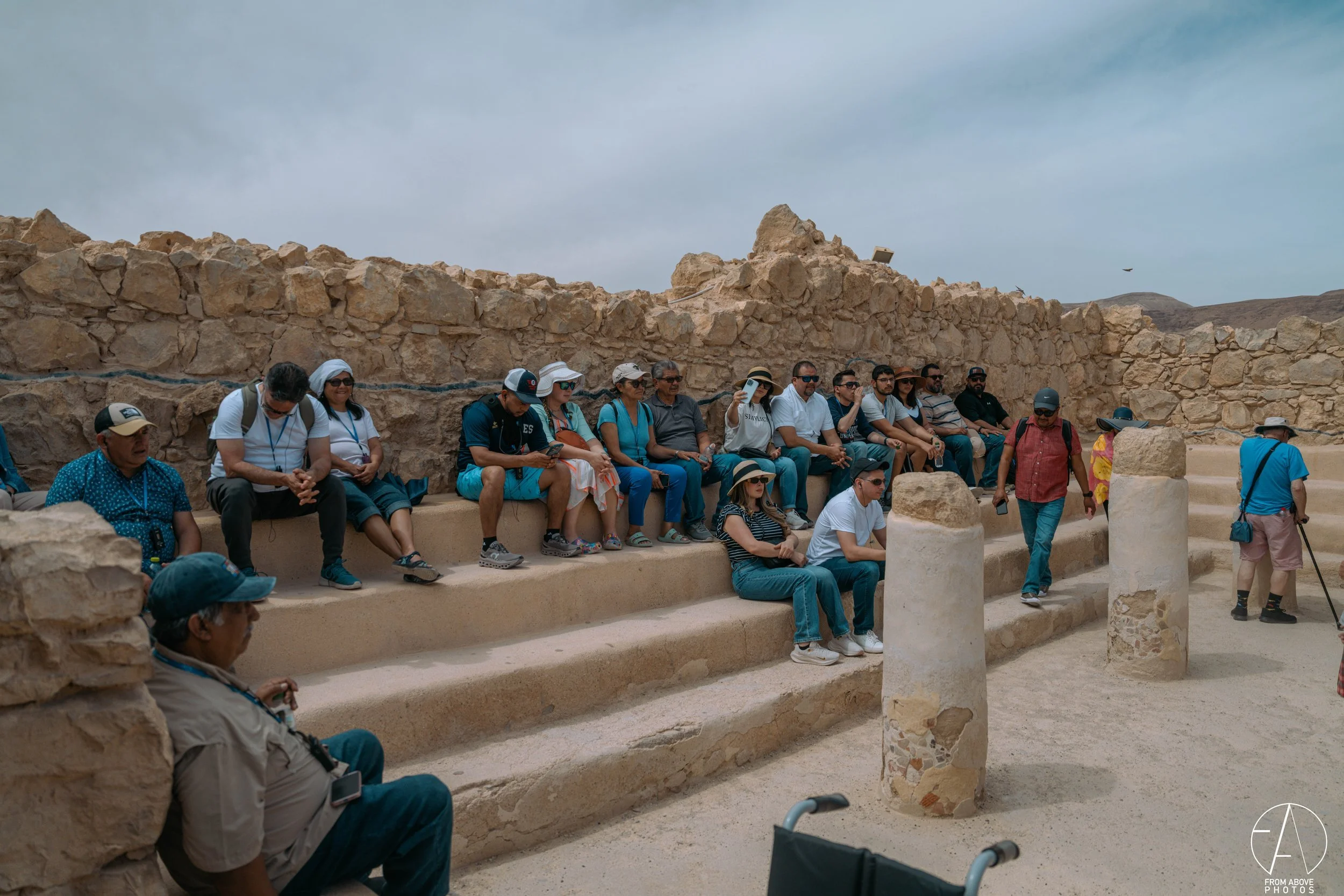 Tourists sitting on stone benches and stairs at an archaeological site, with ruins and a cloudy sky in the background.