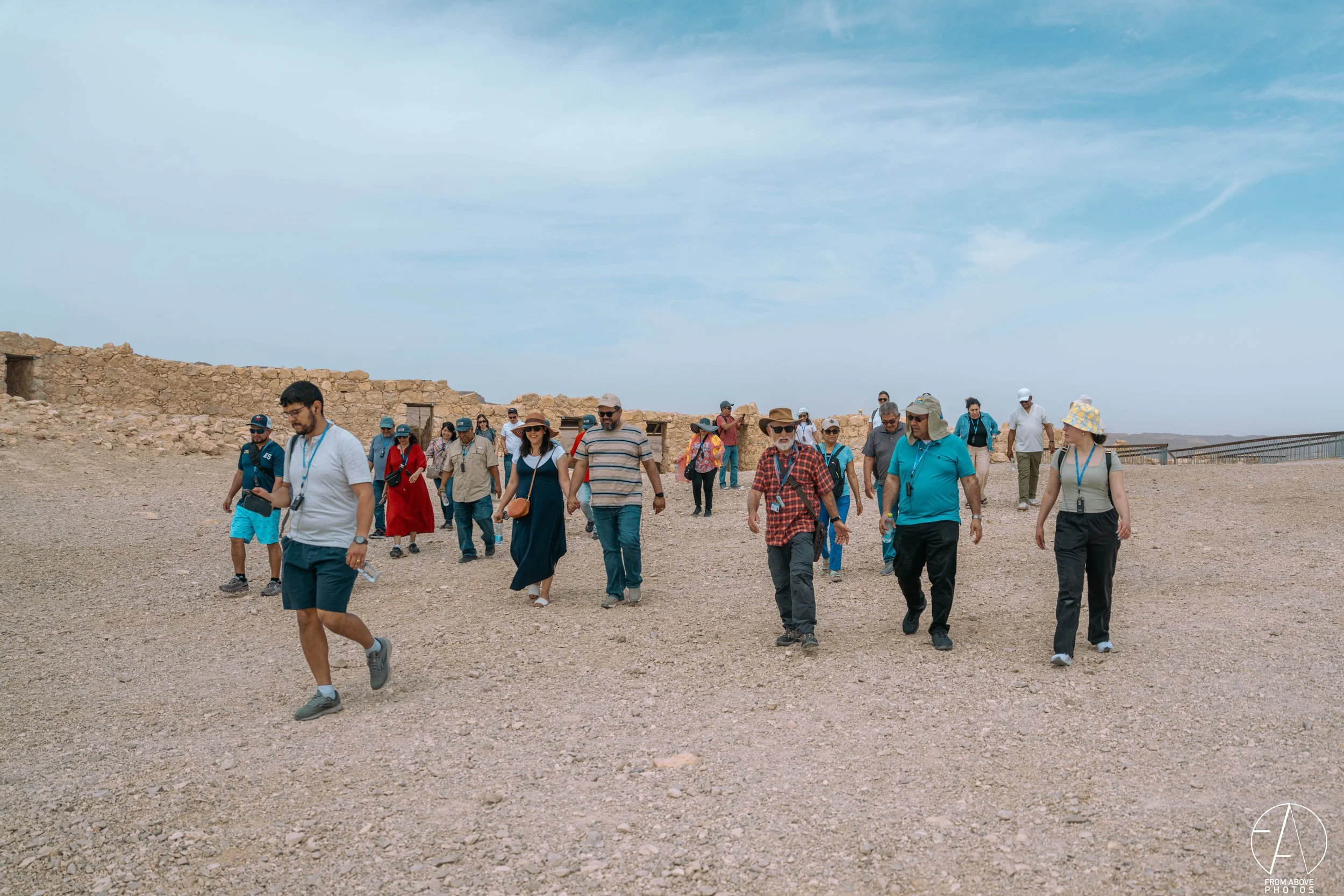 A group of tourists walking outdoors on rocky terrain with stone ruins in the background under a partly cloudy sky.