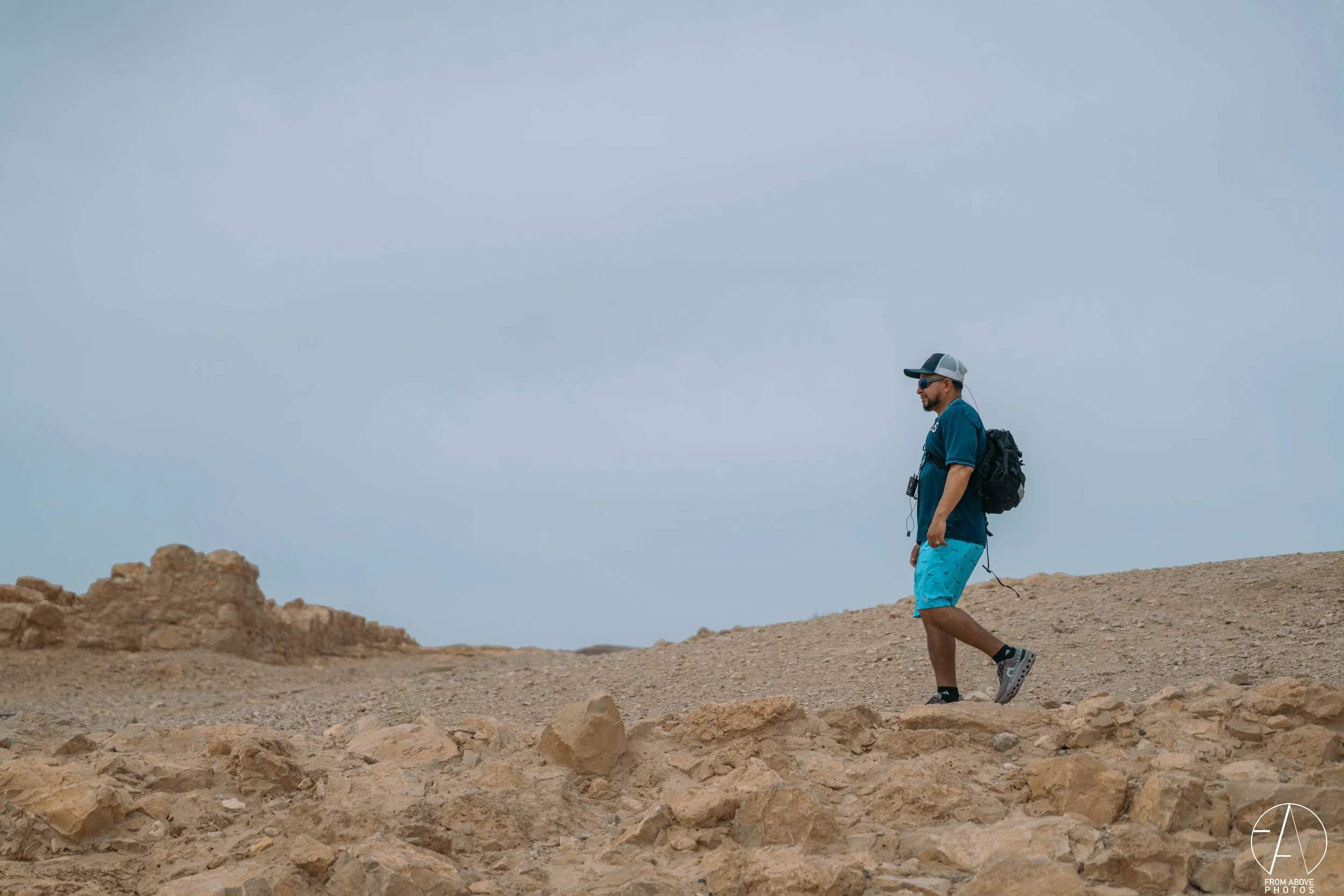 A man wearing a blue t-shirt, blue shorts, a cap, sunglasses, and a backpack walking on a rocky desert landscape under a cloudy sky.