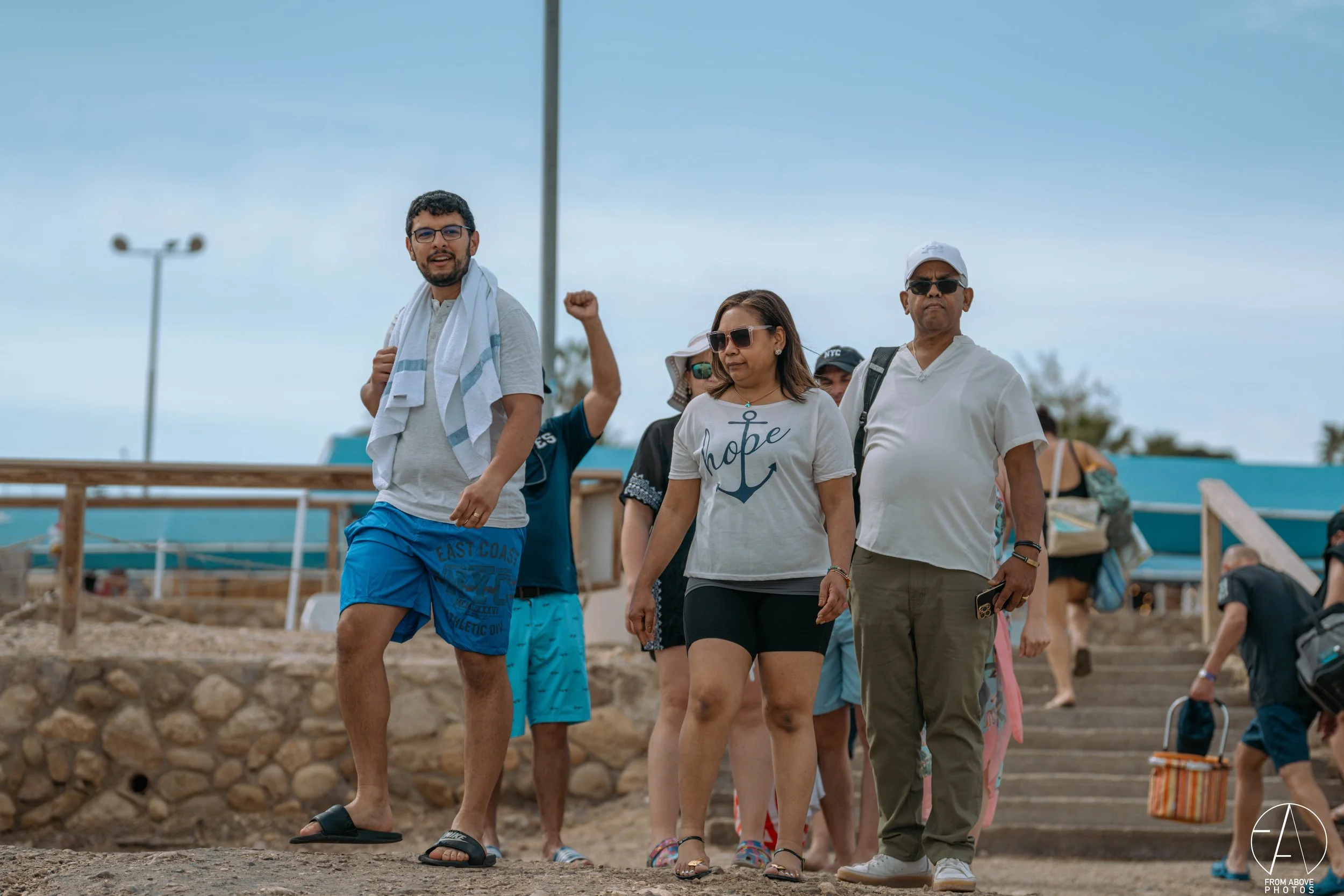 A group of people walking outdoors on a cloudy day, some wearing sunglasses and casual summer clothing. One man is waving with a towel on his shoulder, while others are looking around or using their phones. There are stairs and a wooden railing behin
