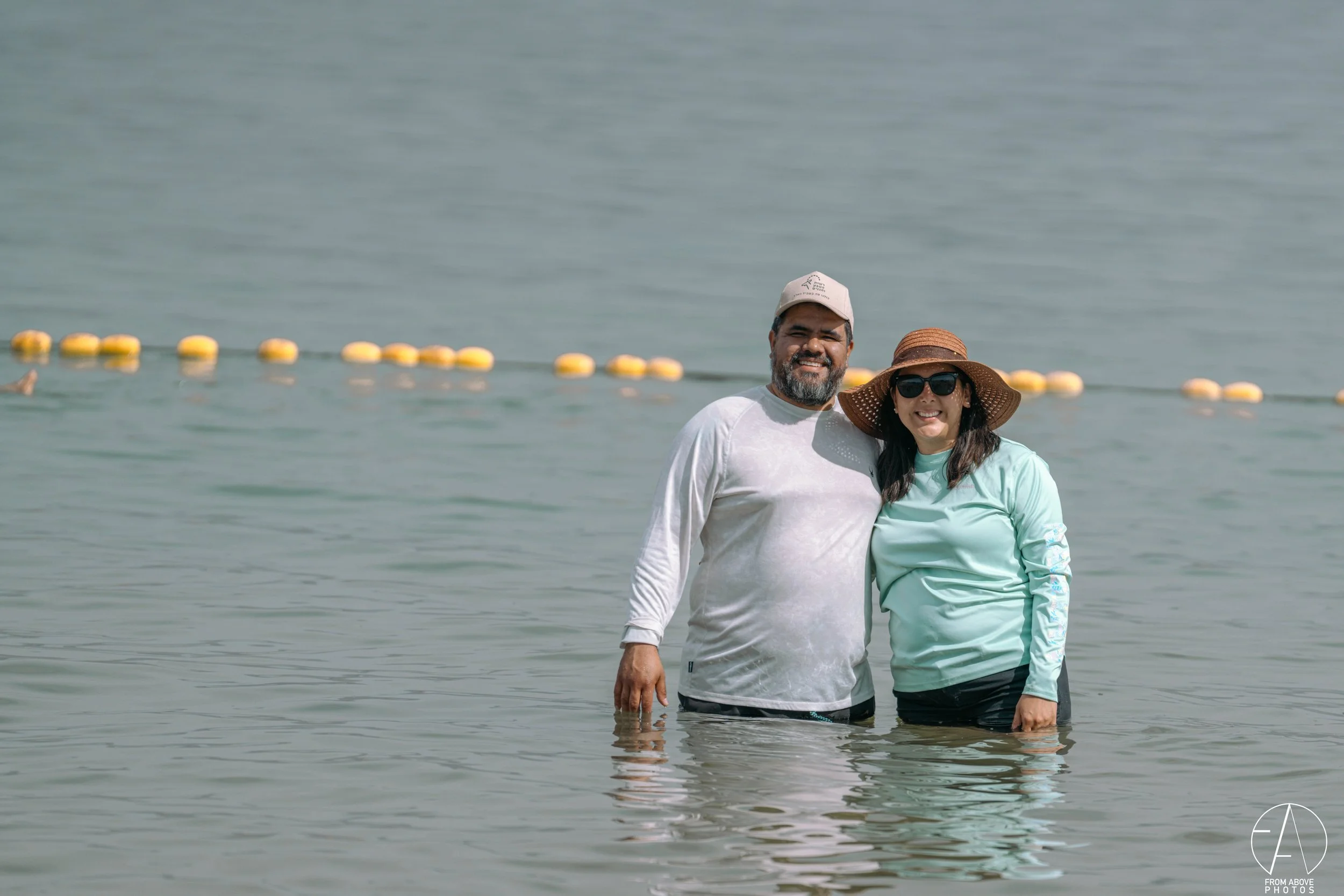 A man and woman standing in shallow ocean water, smiling, with floating yellow buoys behind them, wearing casual beach clothing and accessories.