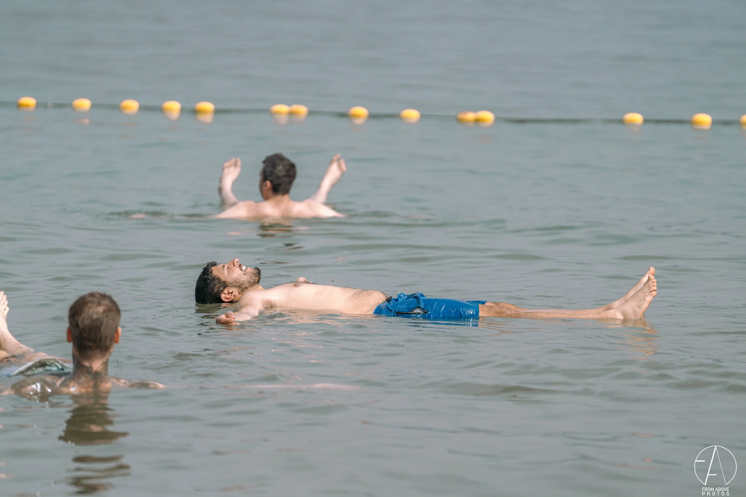 People relaxing and floating in the ocean, with some raising their arms and one person lying on their back with eyes closed. The scene is calm with a line of yellow buoys in the background.