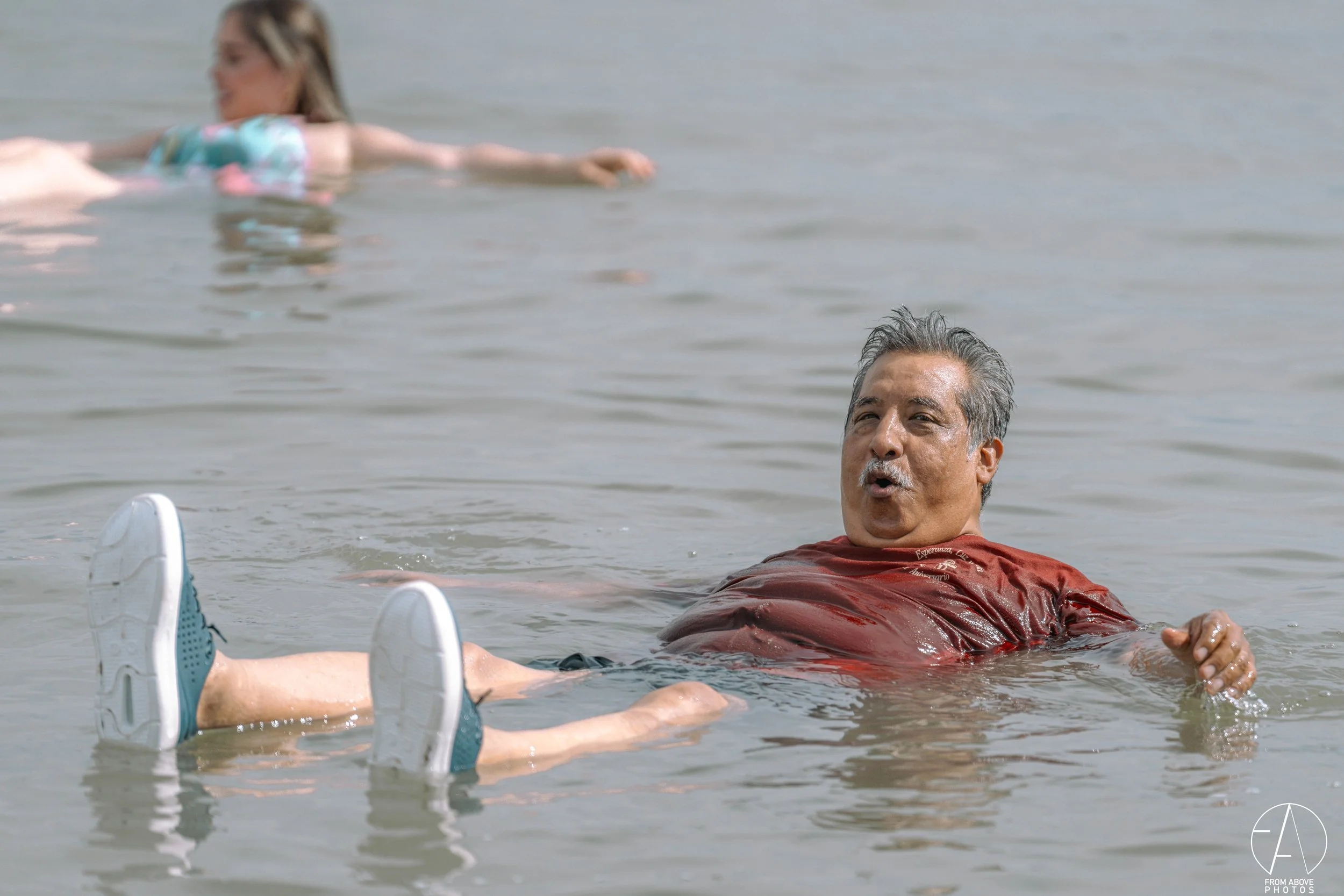 A man with grey hair floating in a body of water, making an expressive face, with a woman swimming in the background.