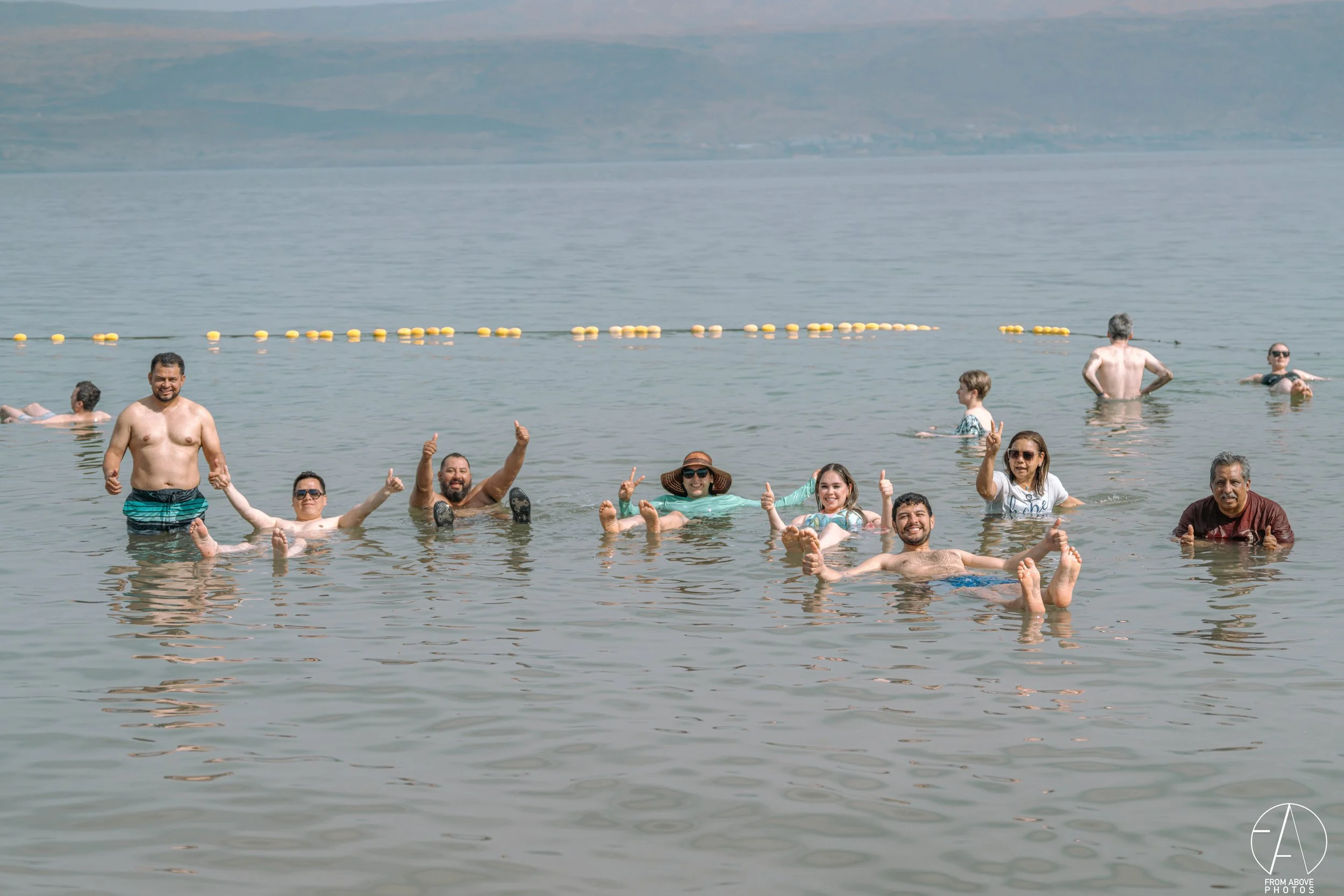 A group of nine people are swimming and floating in a lake, smiling and giving thumbs-up. They are in shallow water with a mountainous landscape in the background.