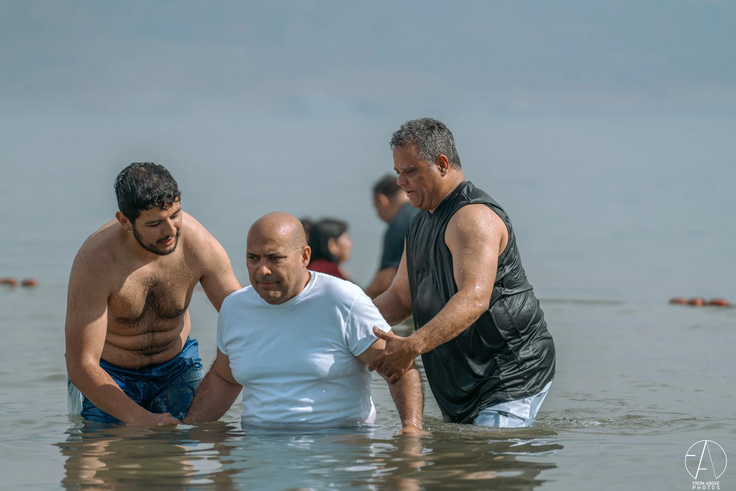 Three men standing knee-deep in water, with one man in a white shirt being assisted by two others, at a beach or lake, during daytime.
