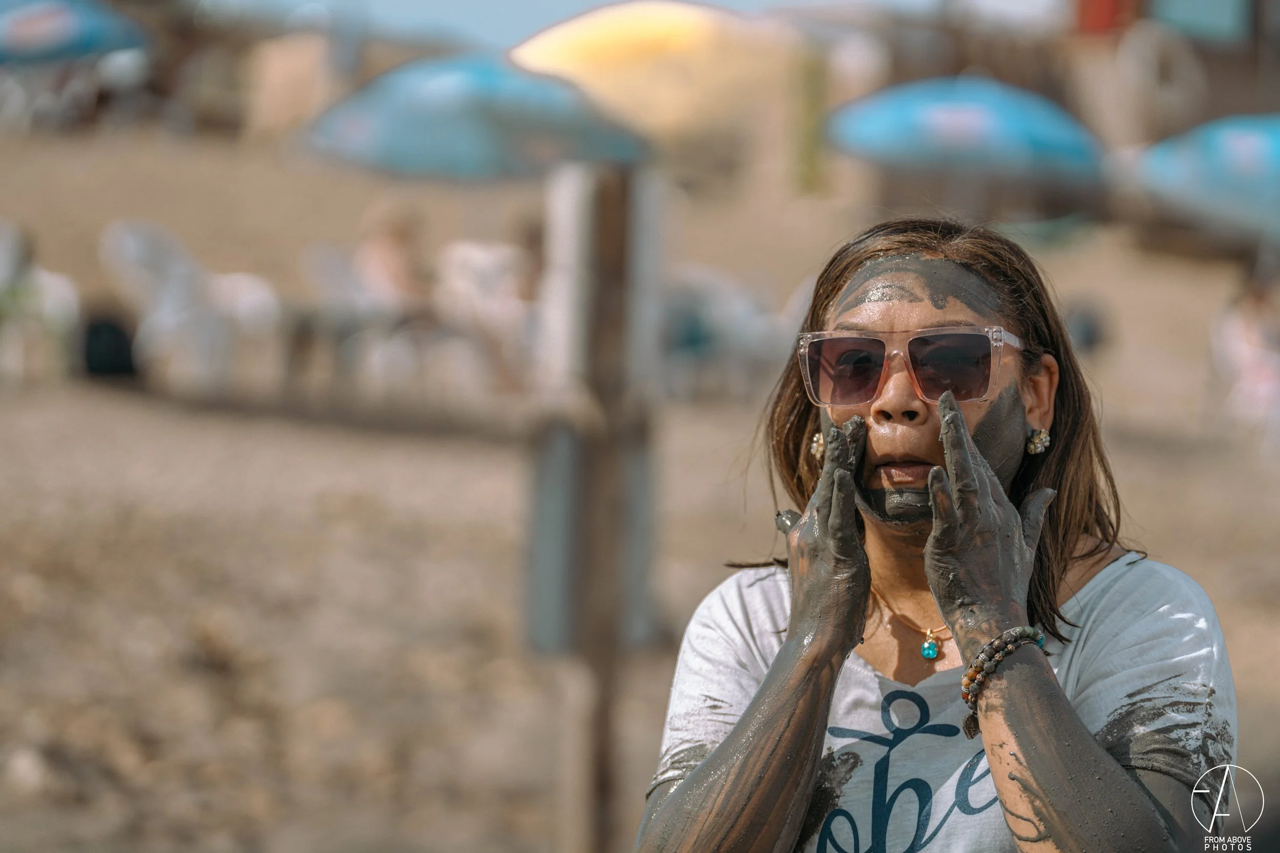 A woman with mud on her face and hands, wearing sunglasses, jewelry, and a white T-shirt, standing outdoors on a beach with umbrellas in the background.