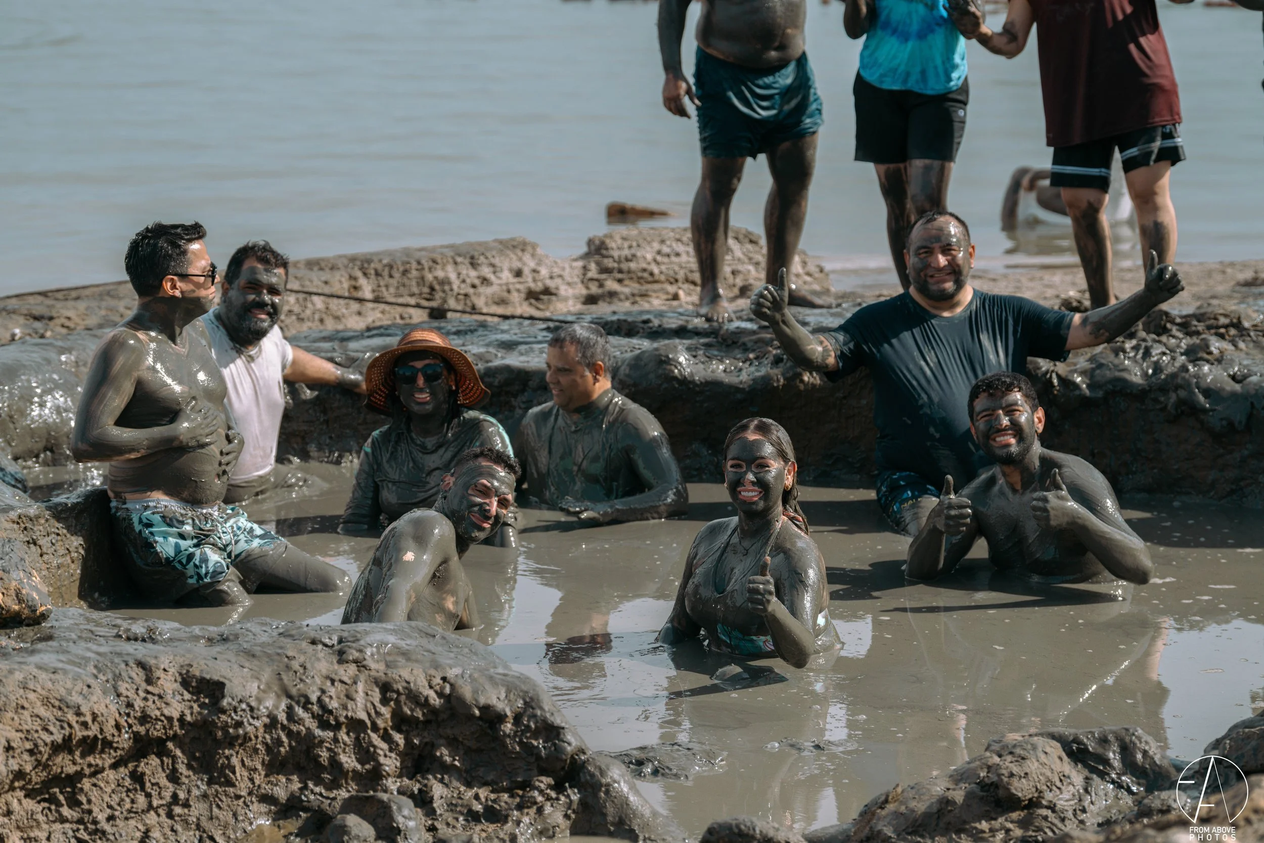 Group of people enjoying in a muddy water pool by the riverbank, some smiling and giving thumbs up, wearing casual clothes and sun hats, with rocks and water in the background.