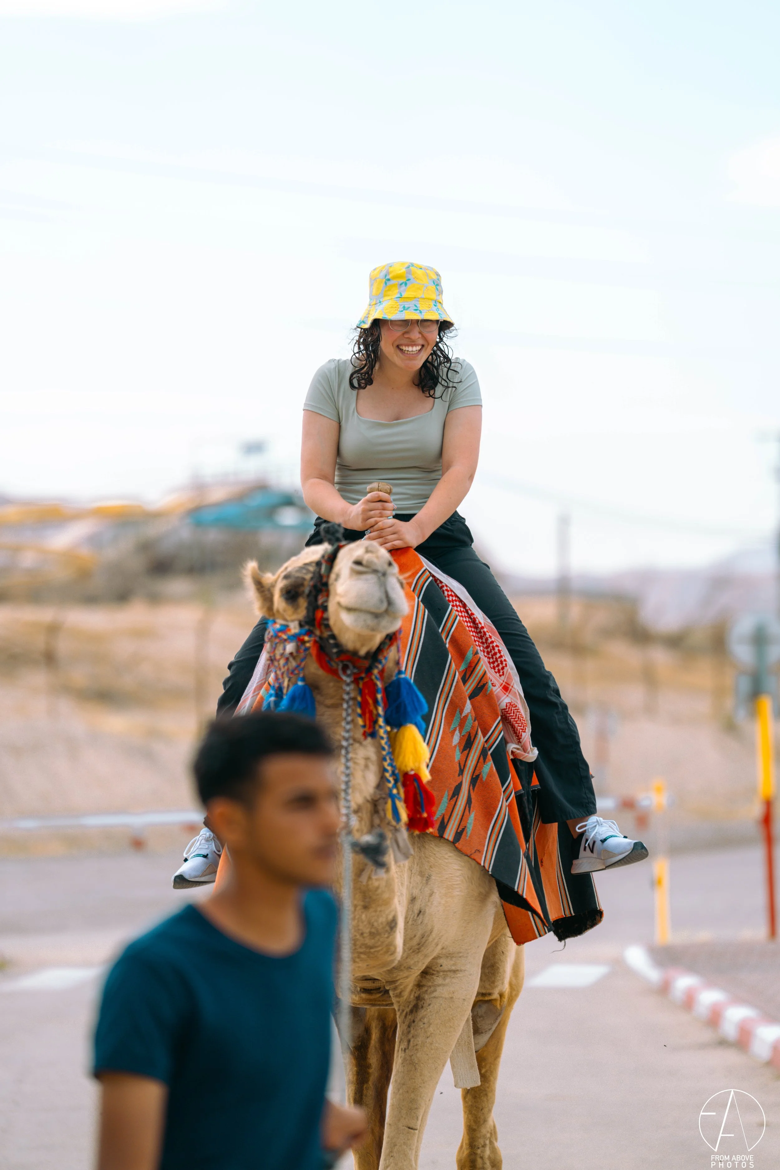 Woman smiling while riding a camel with colorful decorations, wearing a yellow bucket hat, glasses, and a gray t-shirt, in an outdoor setting.