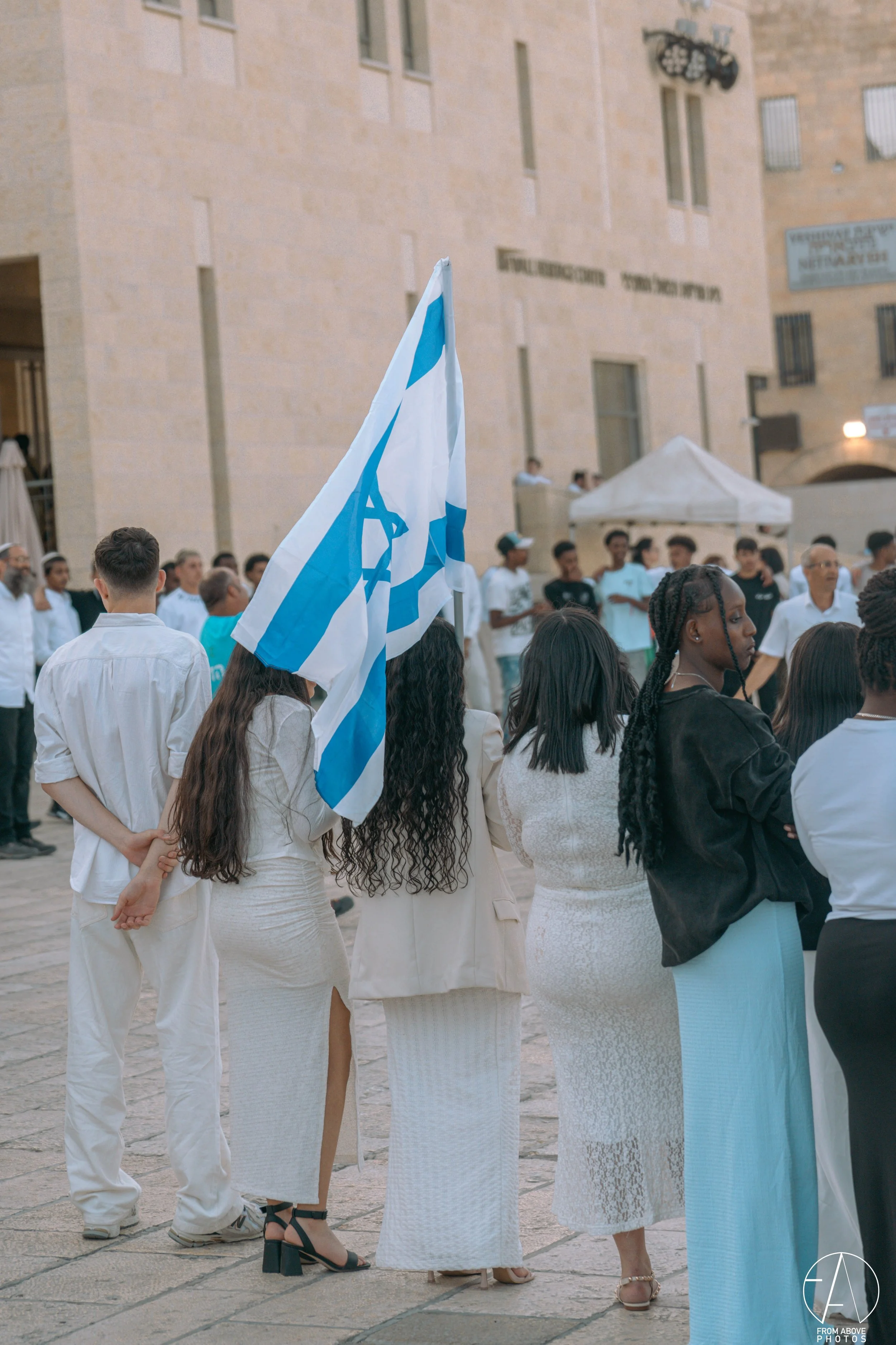A group of diverse women standing with their backs to the camera, one holding an Israeli flag, in an outdoor urban setting with other people and buildings in the background.