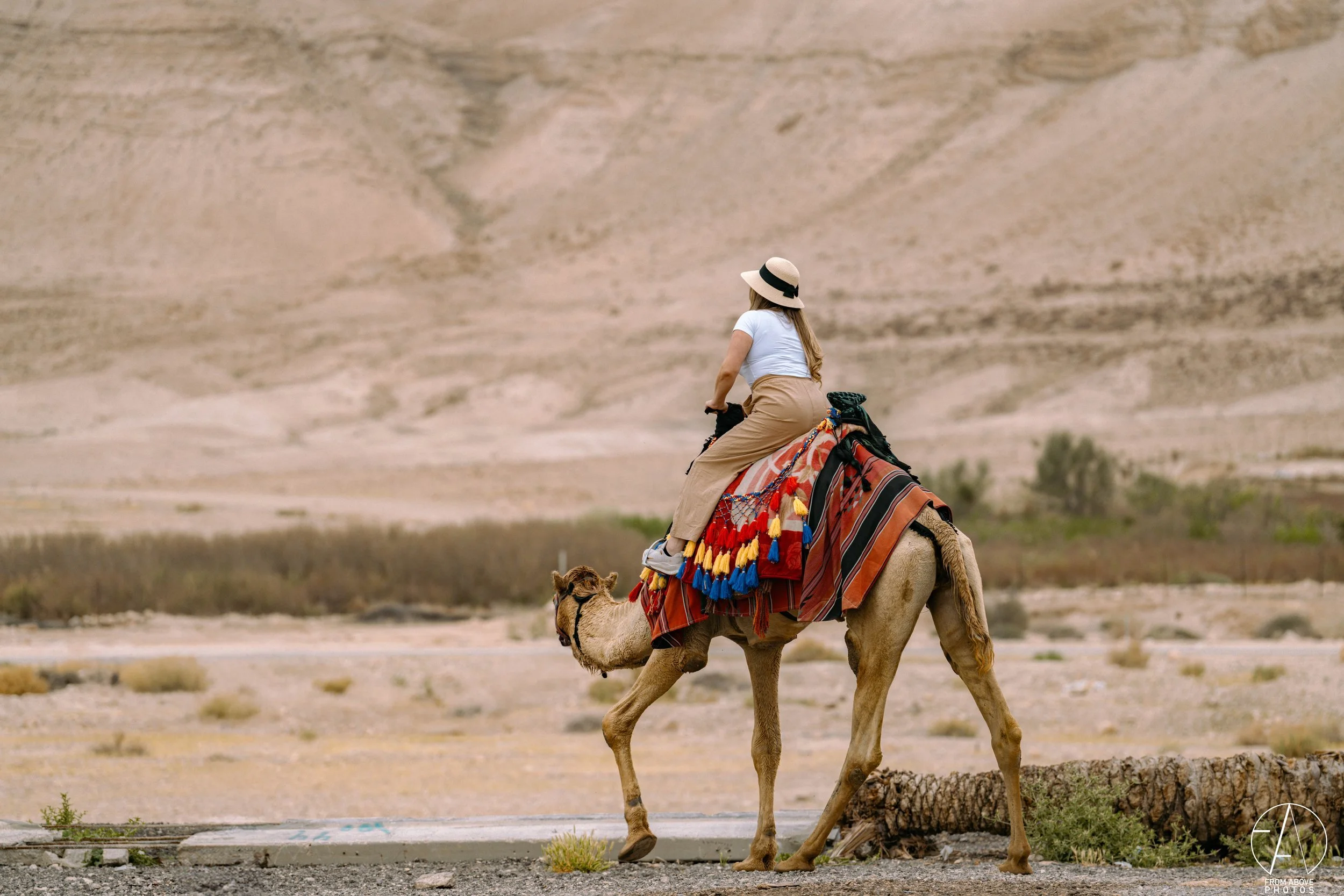 A woman riding a camel in a desert landscape with mountains in the background, wearing a wide-brimmed hat, white t-shirt, and beige pants.