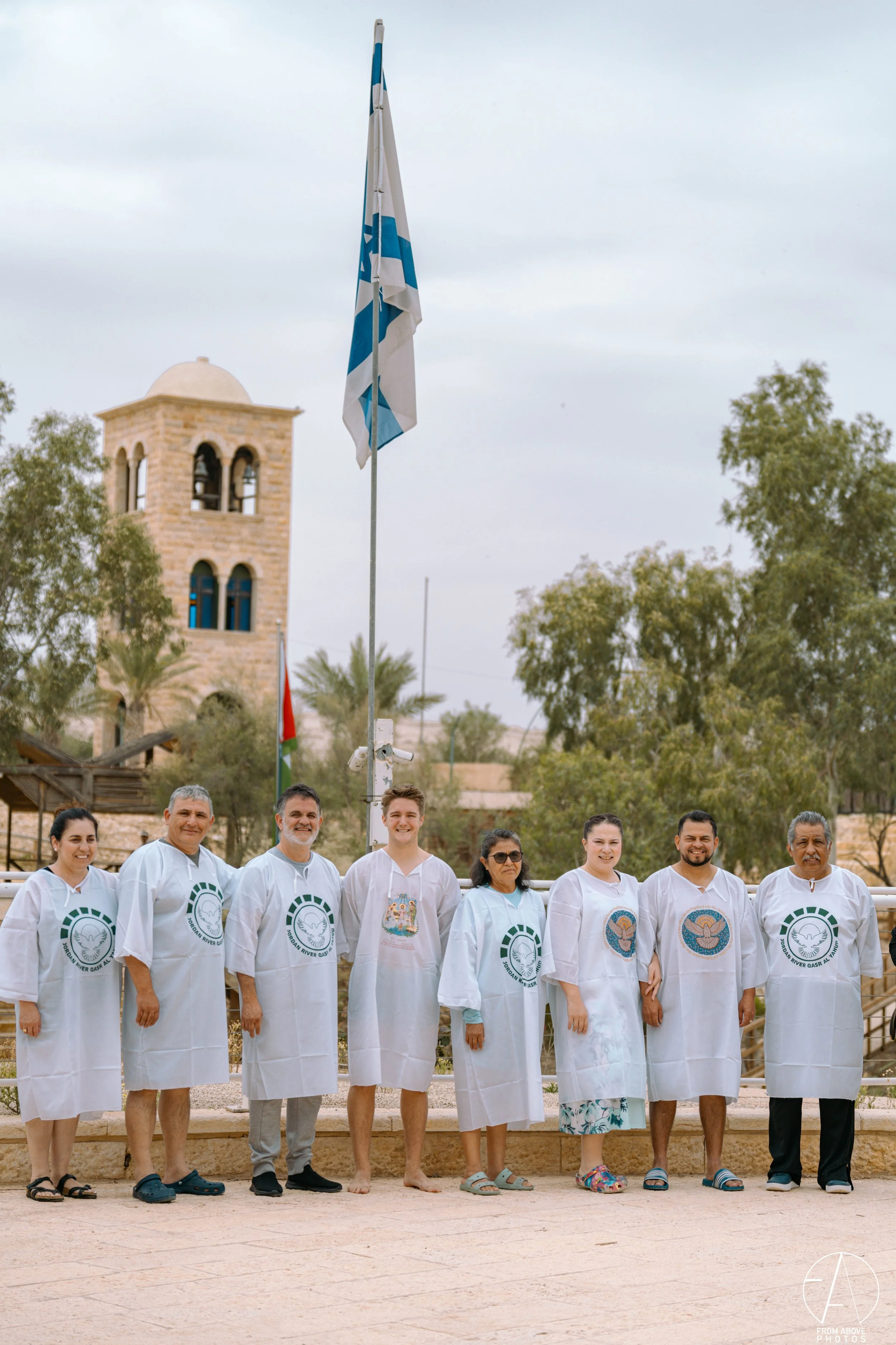 A group of nine people standing in a line in front of a flagpole with a Greek flag, wearing white smocks with various logos and symbols, with ancient architecture and trees in the background.