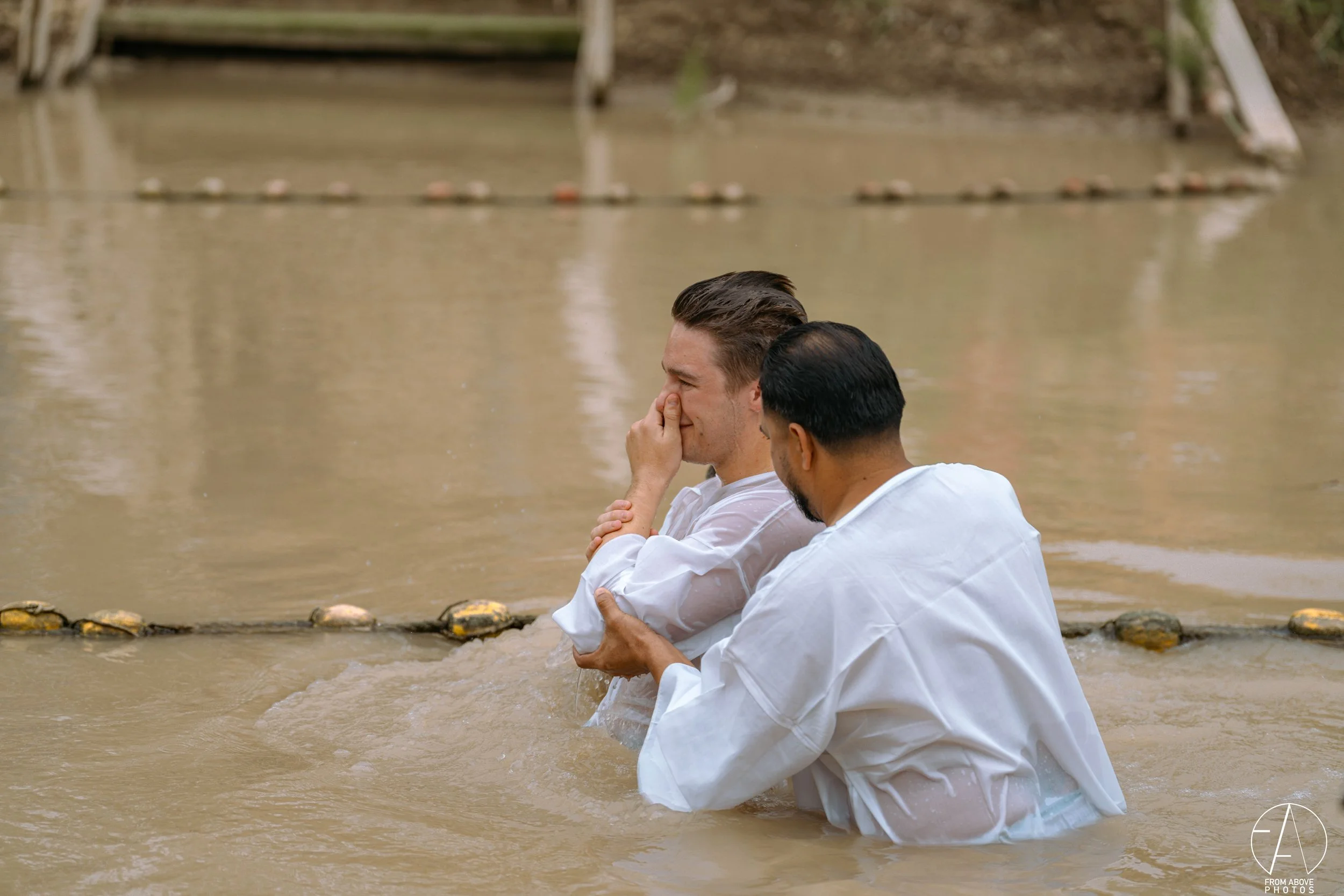 Two men in white shirts baptizing each other in a muddy river, with a dock in the background.