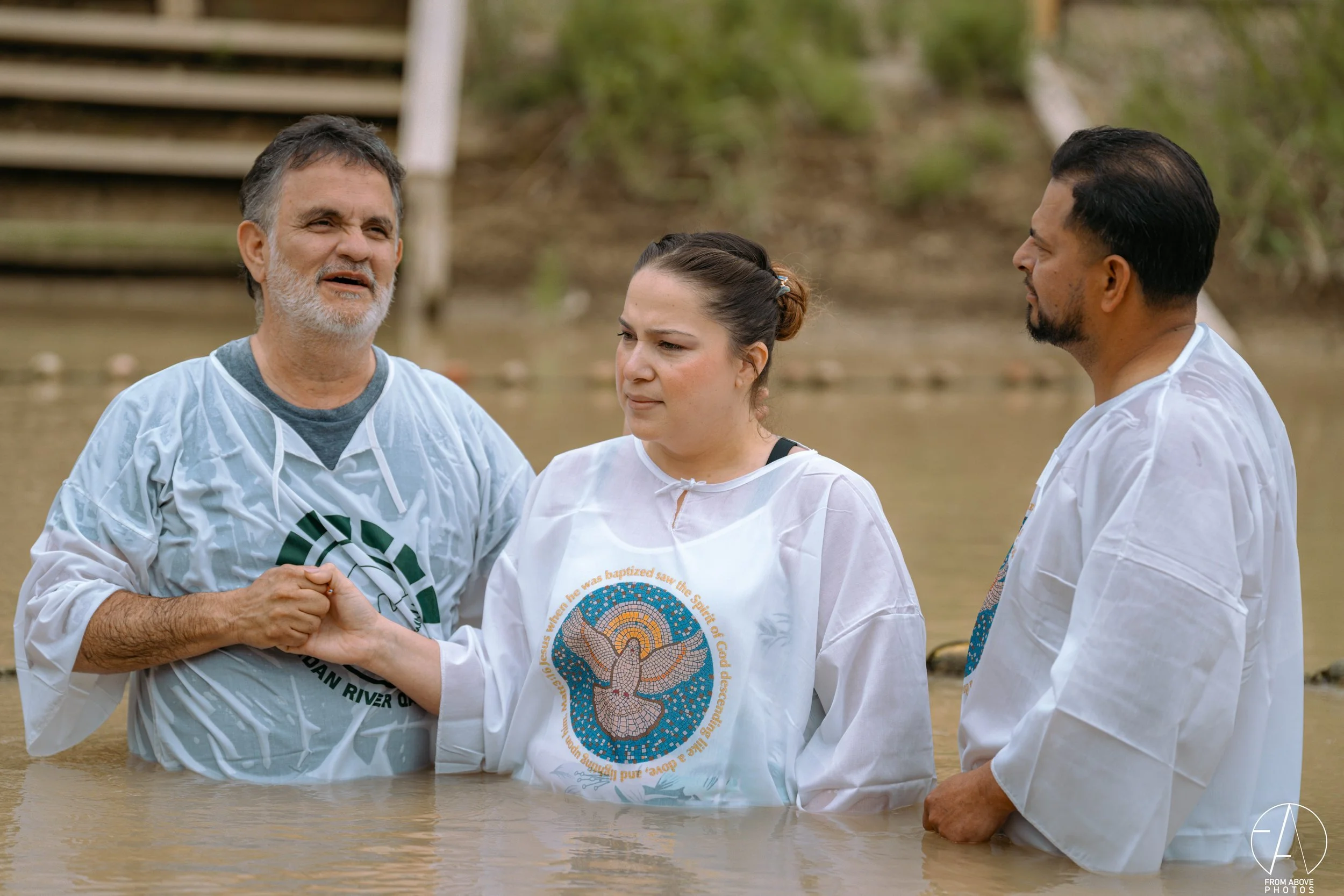 A couple of people baptized in a river during a religious ceremony, with two men and a woman standing in water, holding hands, with a wooden staircase in the background.