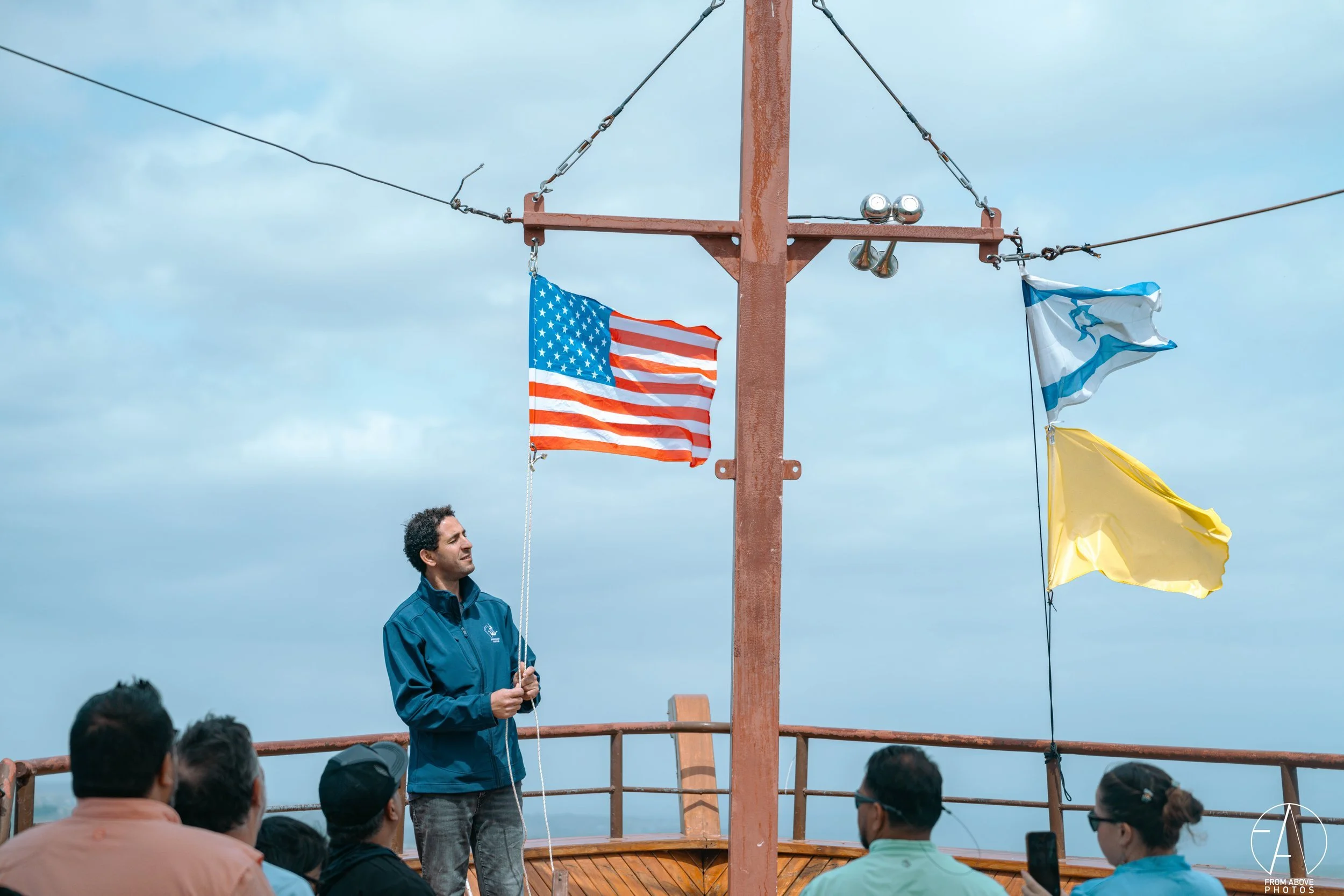 A man speaking to a group of people outdoors on a boat deck, with the American flag, a blue and white flag, and a yellow flag hanging from a mast.
