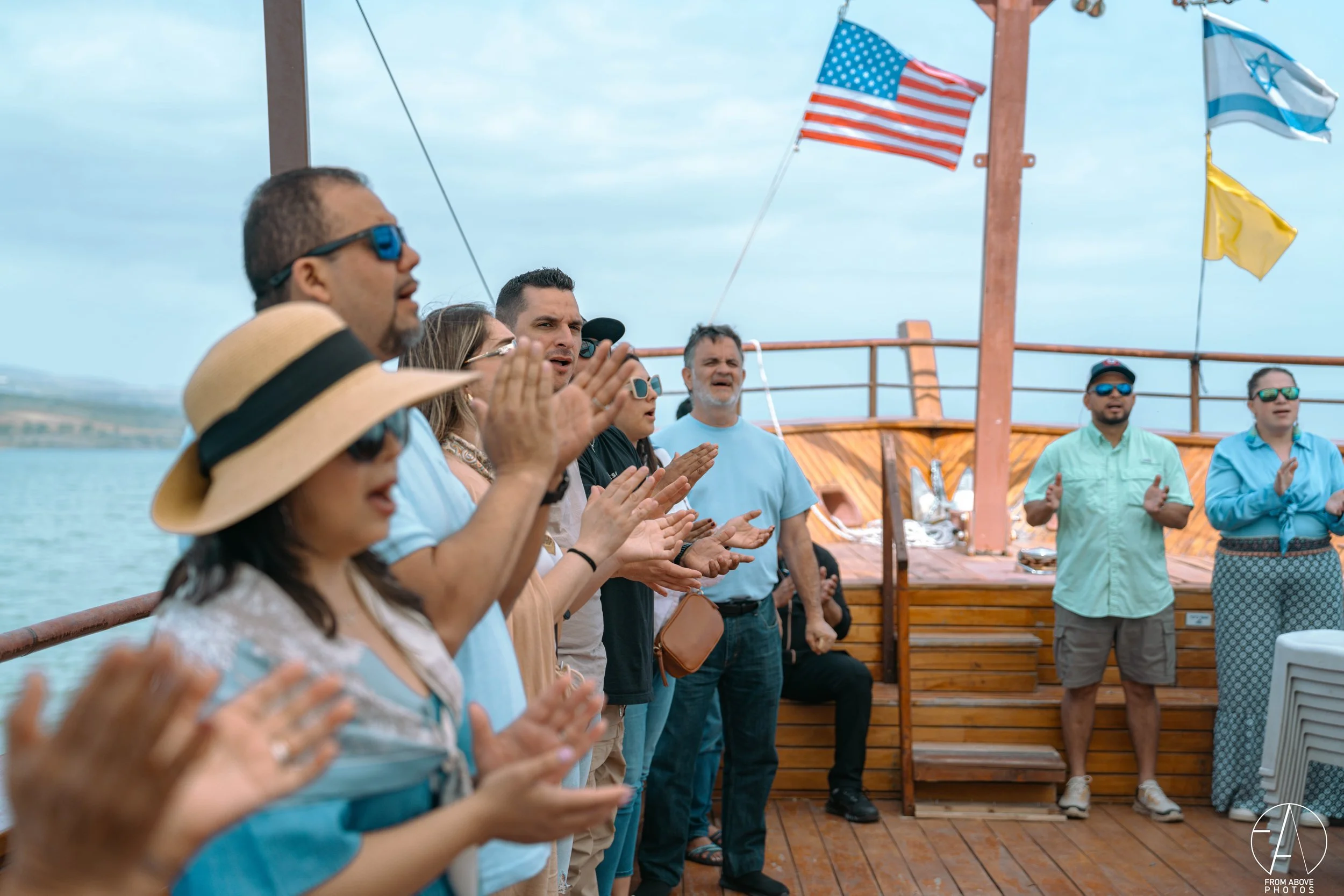 A group of people on a boat deck, participating in a prayer or blessing, with flags of the United States, Israel, and Ukraine flying in the background.