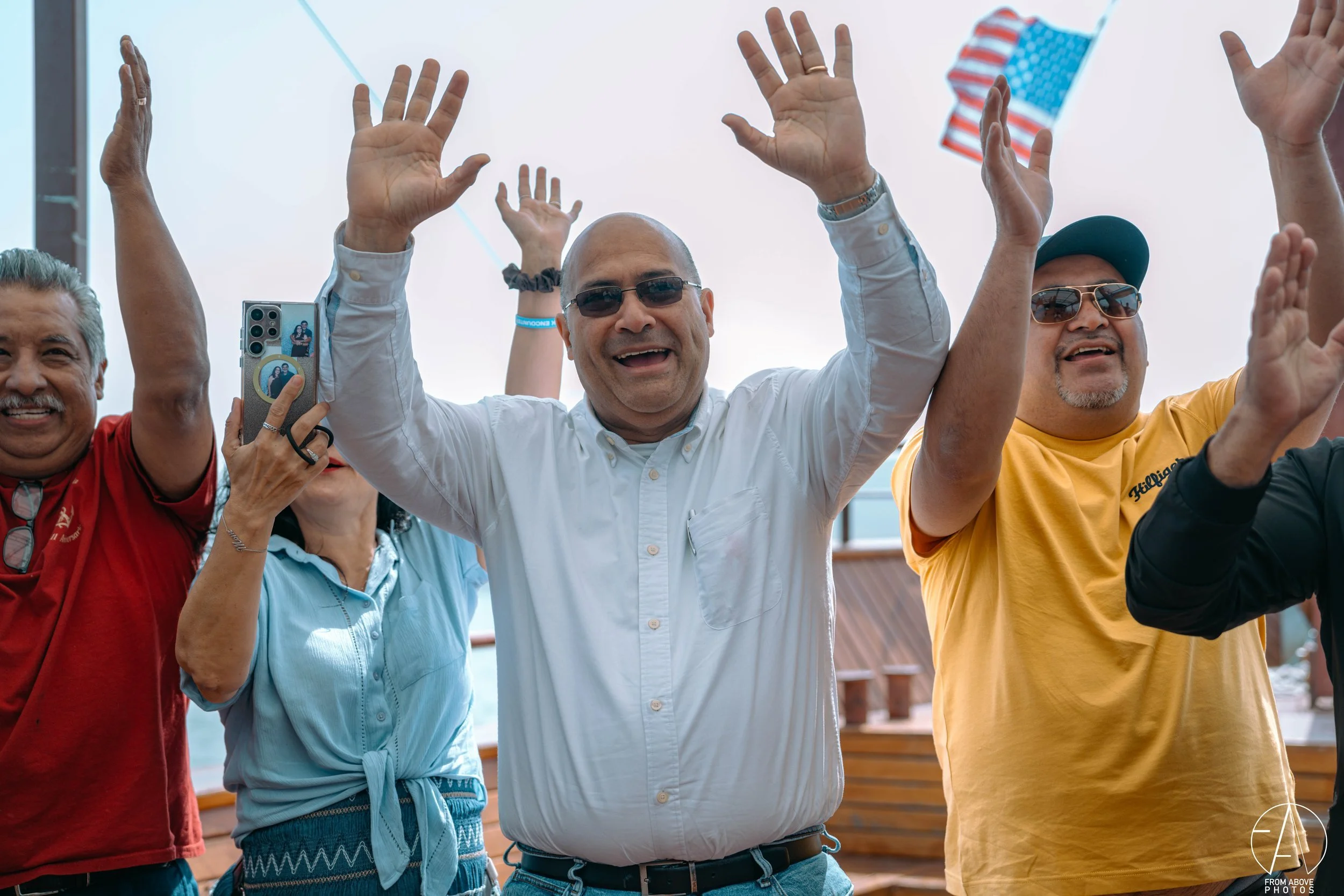 A group of people on a boat deck cheering and raising their hands, with some wearing sunglasses and casual summer clothing