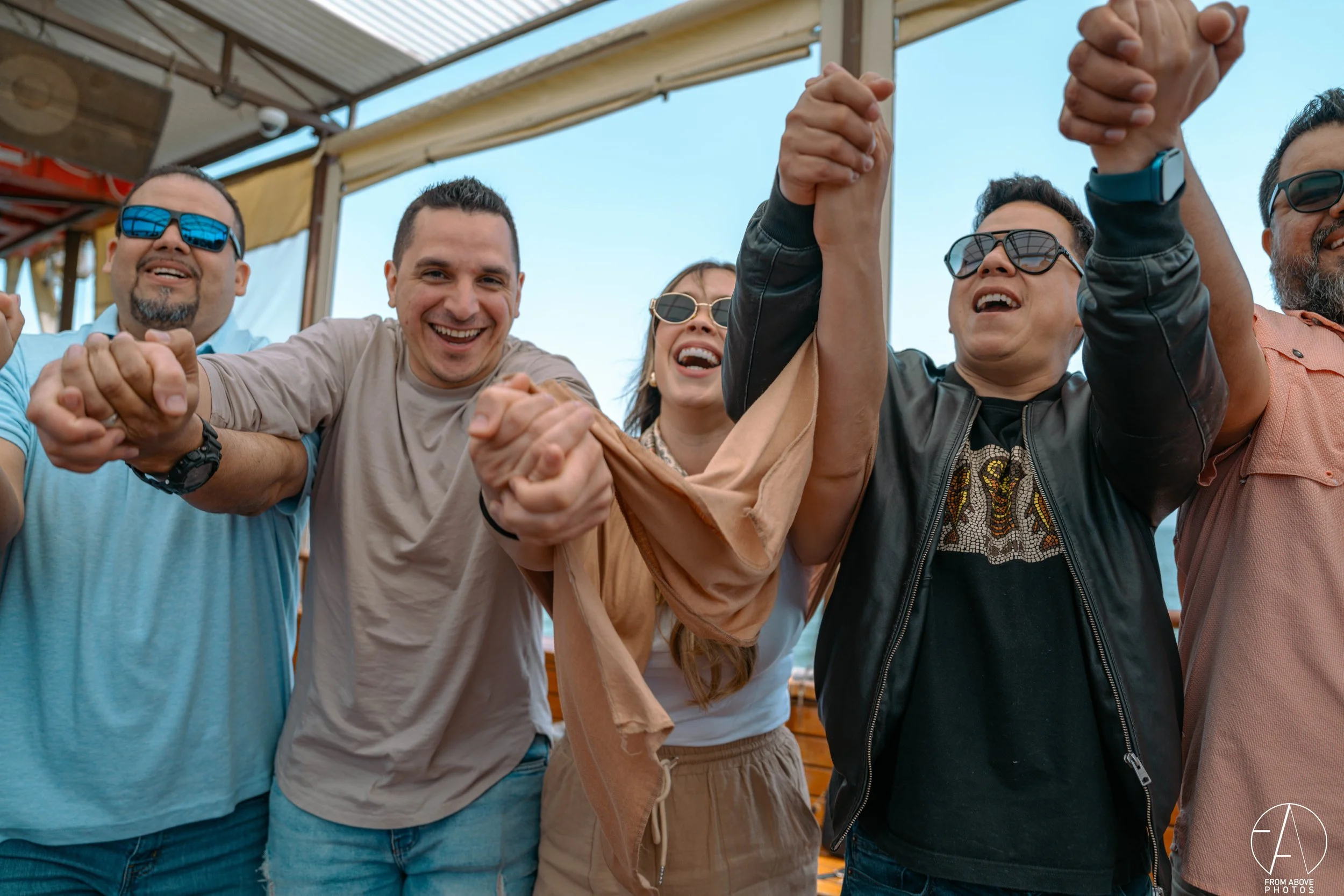 A group of diverse smiling people holding hands and celebrating outdoors under a canopy, wearing casual clothing and sunglasses.