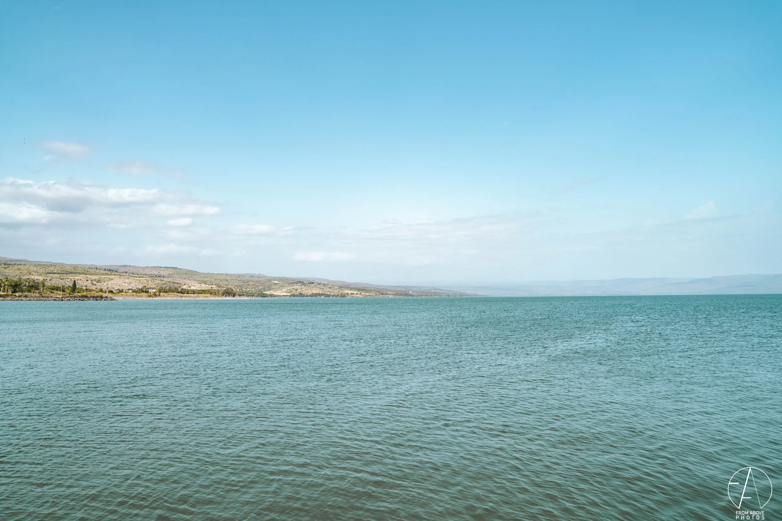 A calm body of water extending to the horizon, with a shoreline and rolling hills in the distance under a clear blue sky with a few scattered clouds.