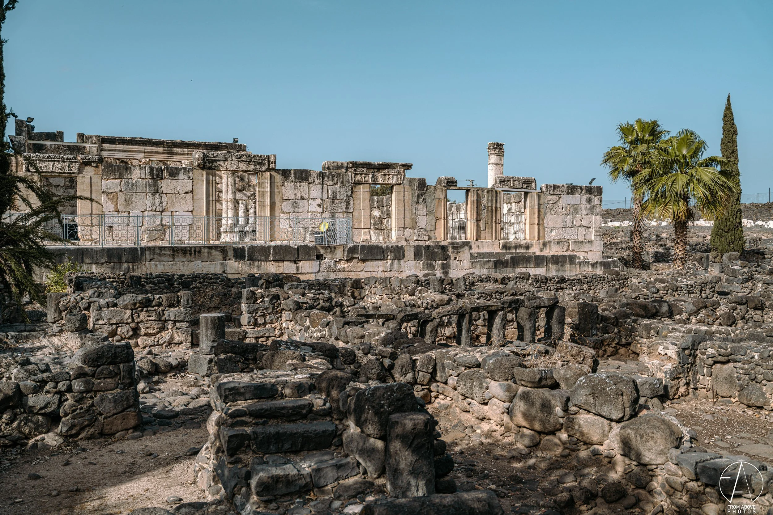 Ancient stone ruins with scattered black rocks and partial stone walls, palm trees, and clear blue sky in the background.