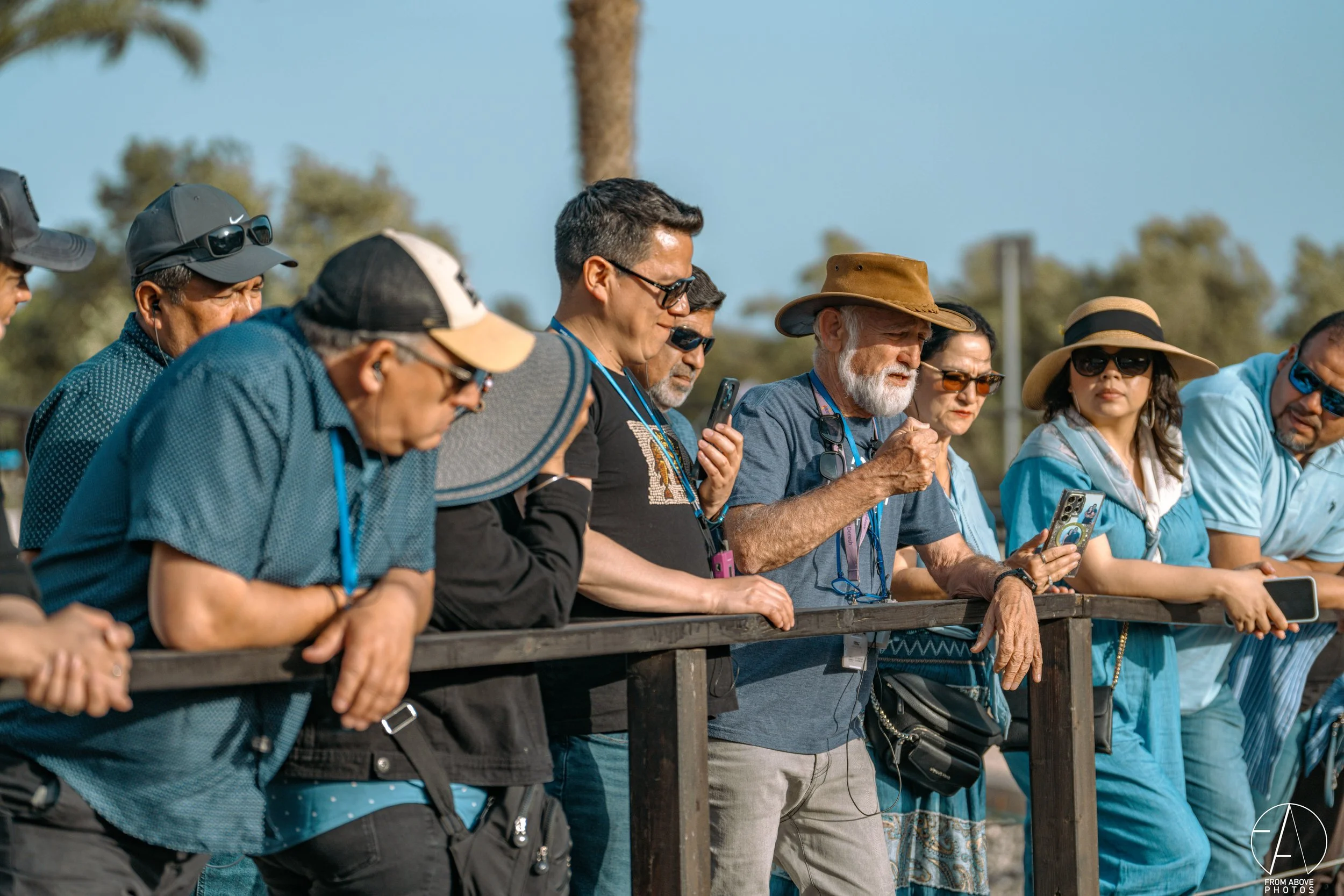 A diverse group of people, including men and women of different ages, are standing behind a wooden railing outdoors, watching something. Some are wearing hats, sunglasses, and lanyards, and many are holding smartphones.