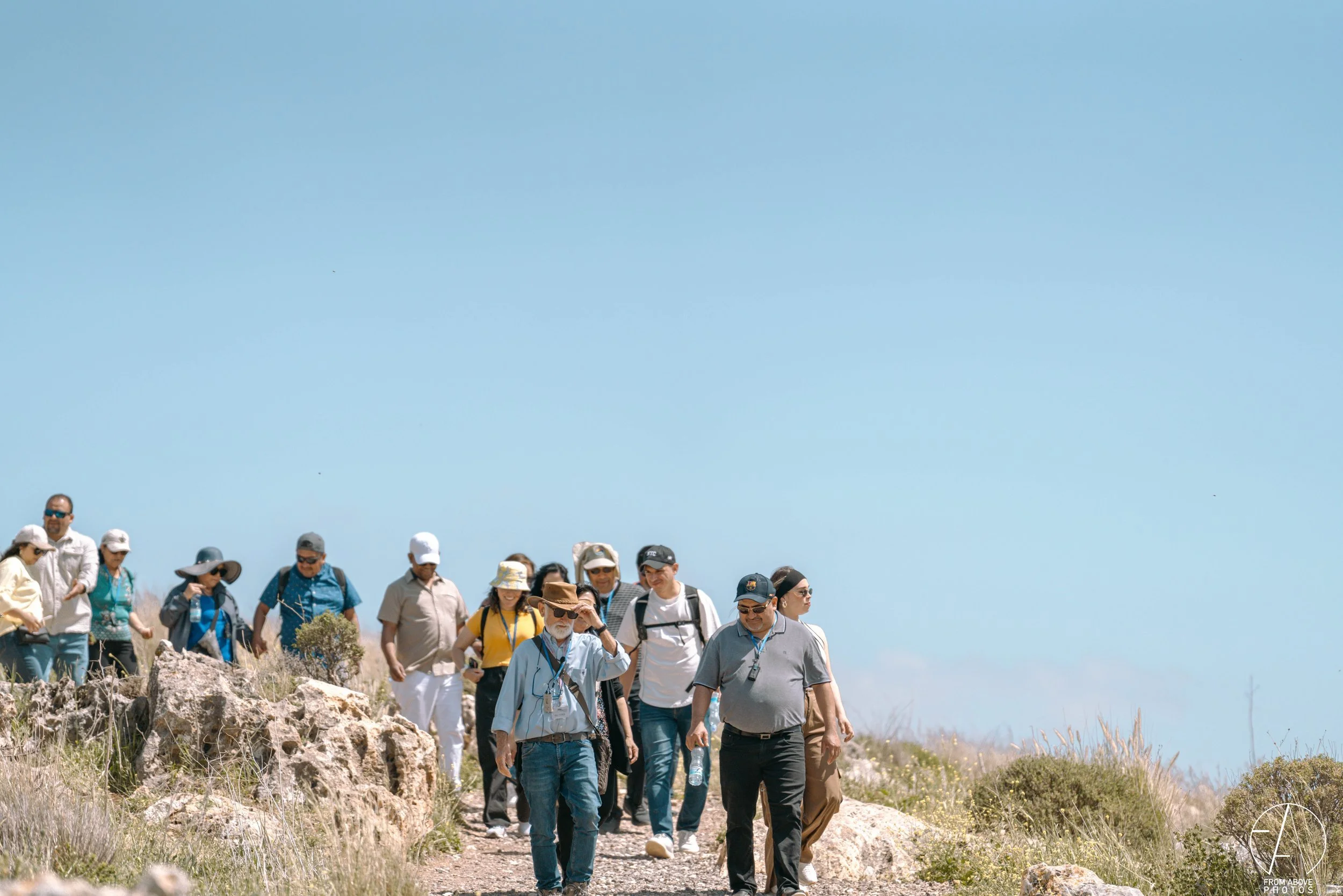 A group of people hiking on a rocky trail in a sunny, open landscape with sparse vegetation and a clear blue sky.