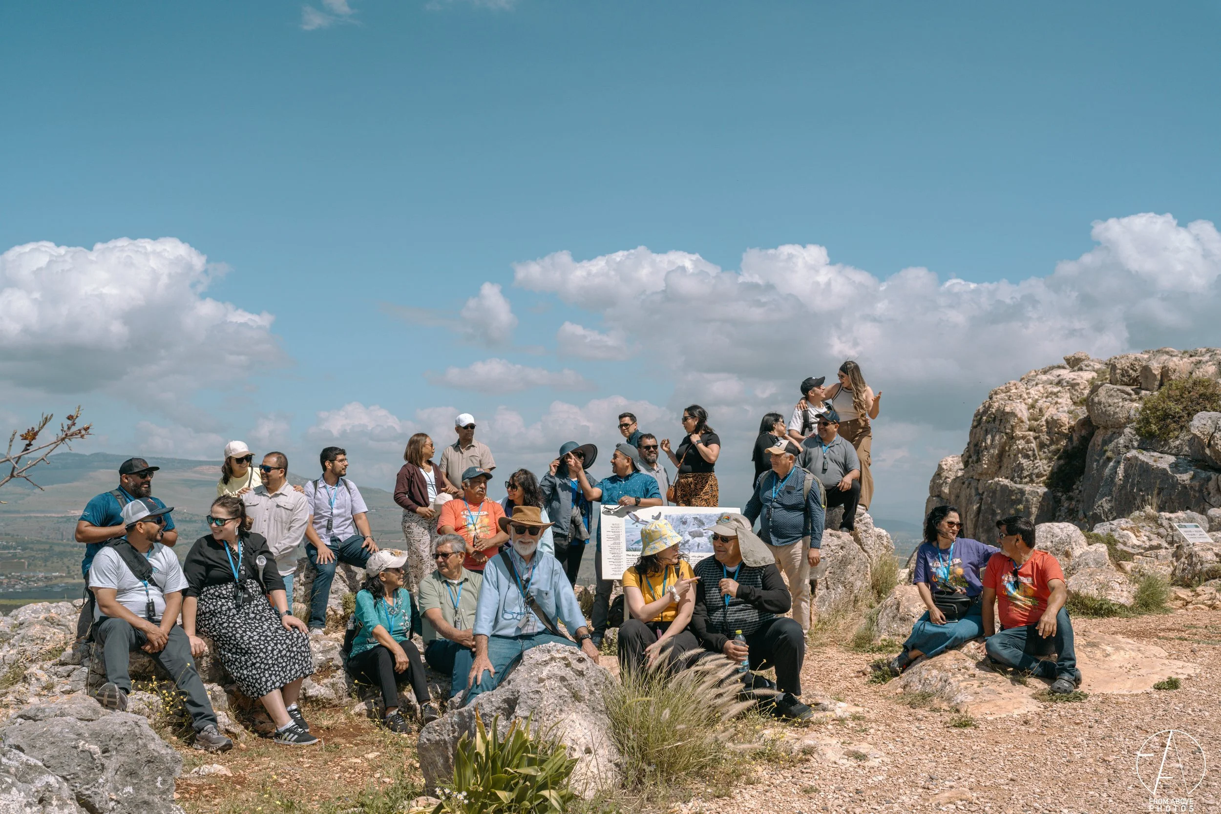 Group of people gathered outdoors on large rocks in a scenic, mountainous landscape with a blue sky and scattered clouds, some sitting and some standing, many wearing hats and sunglasses.