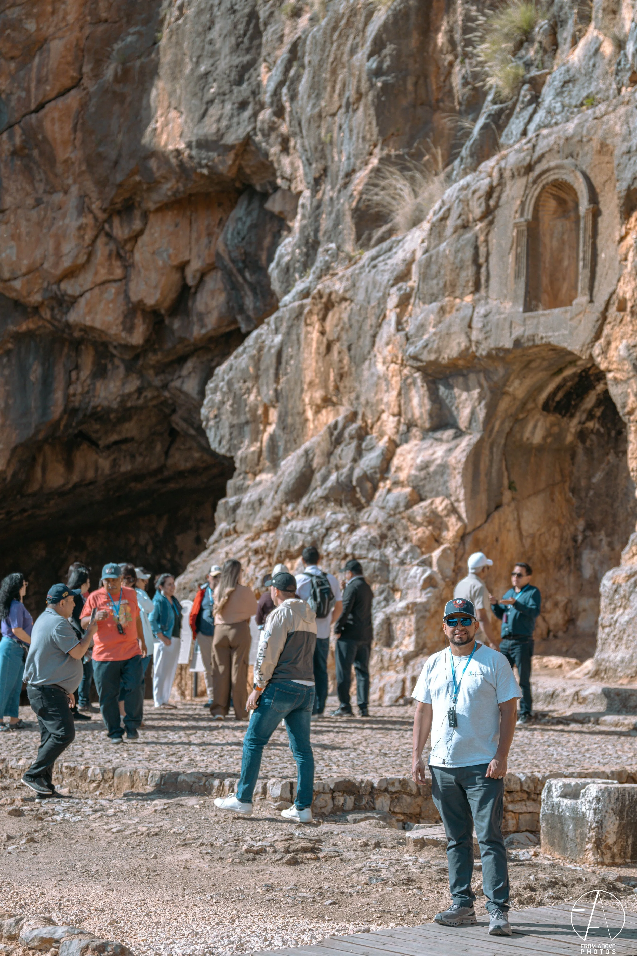 Tourists visiting an archaeological site with ancient rock carvings on a cliff face, with some people taking photos and others observing.