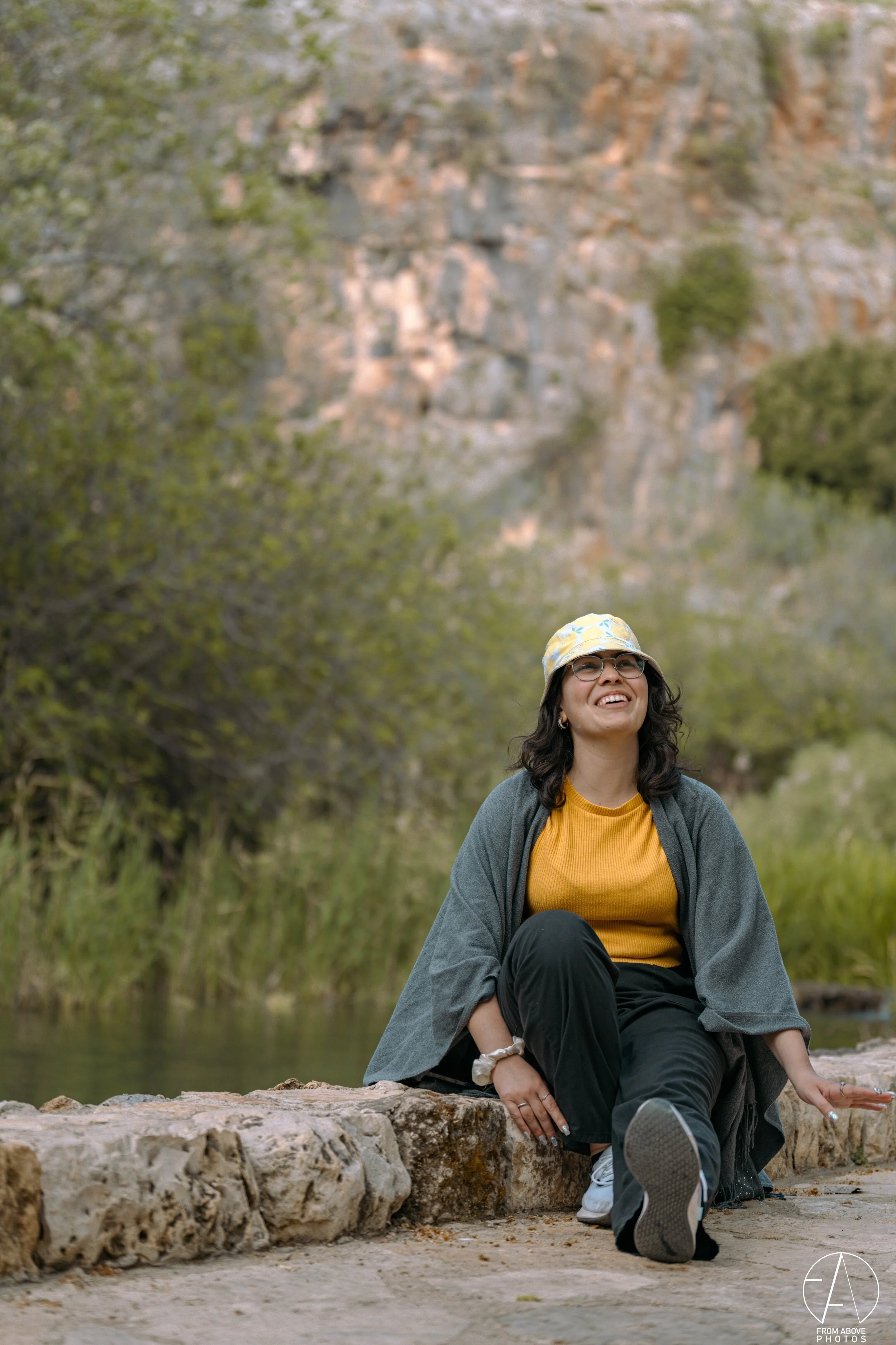 A woman sitting on a stone ledge outdoors, smiling and looking up. She is wearing glasses, a yellow shirt, a gray cardigan, black pants, and a hat with a yellow pattern. There is greenery and a rocky cliff in the background.