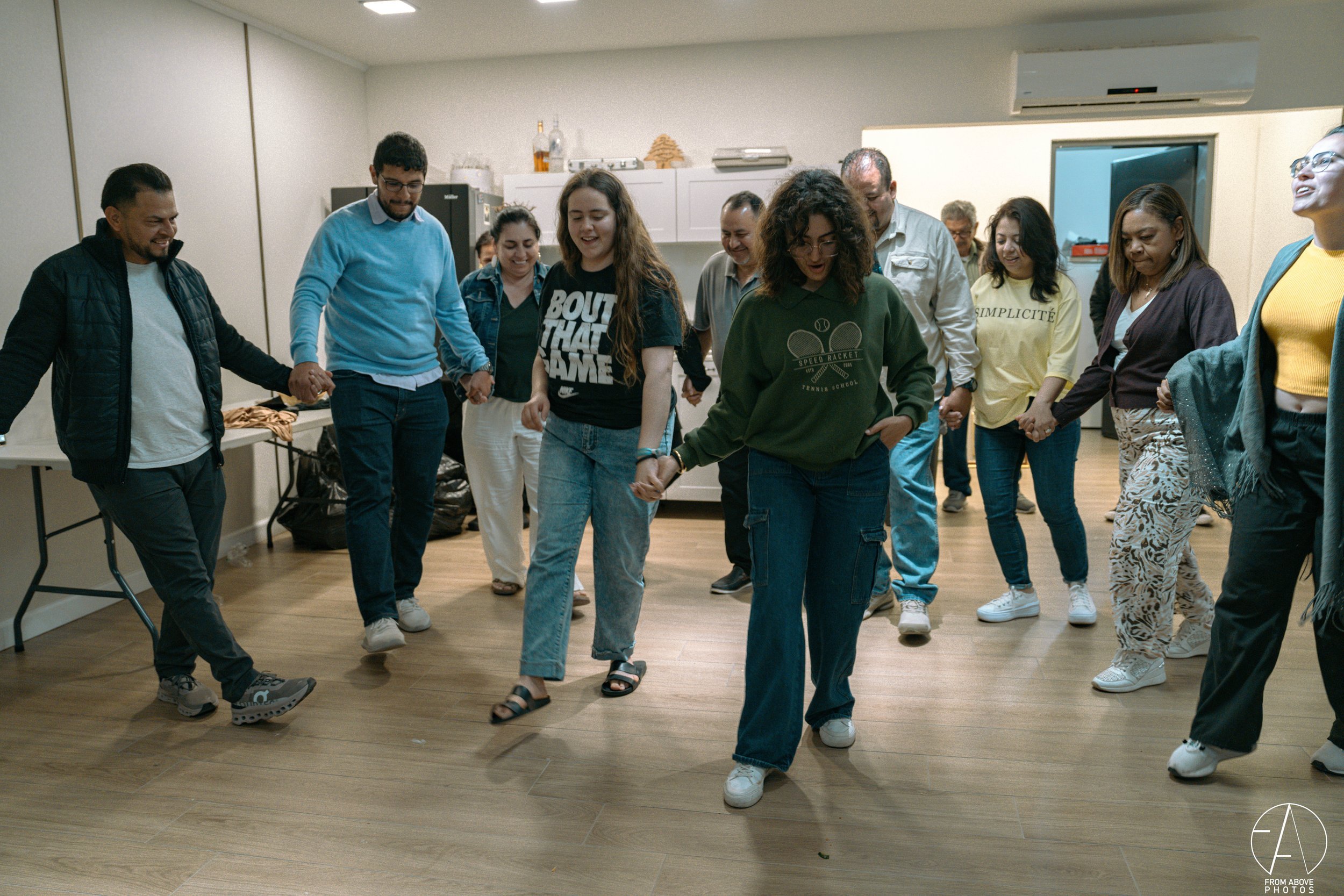 A group of diverse people holding hands and dancing in a circle in a room with wooden floors and a white wall.