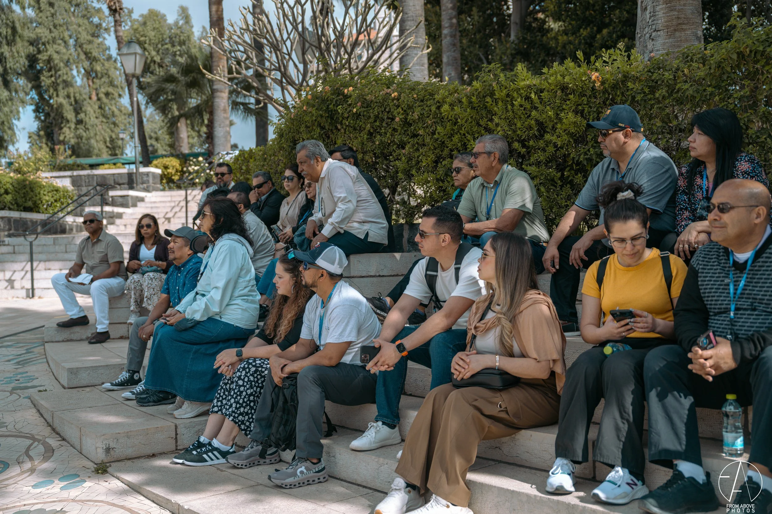 Group of people sitting on stone steps outdoors, some looking at their phones, others talking, with trees and bushes in the background.