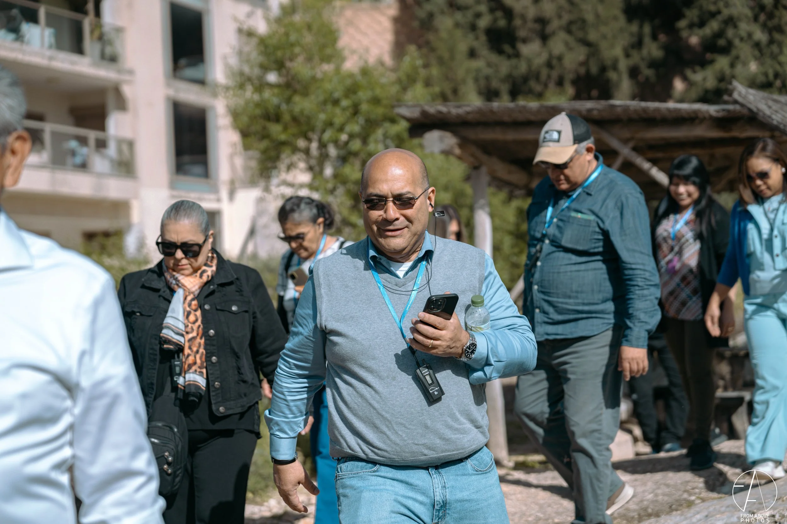 A group of people walking outdoors, some wearing sunglasses and lanyards, with a man in the front using a cellphone and holding a water bottle, in a park-like setting with trees and buildings.